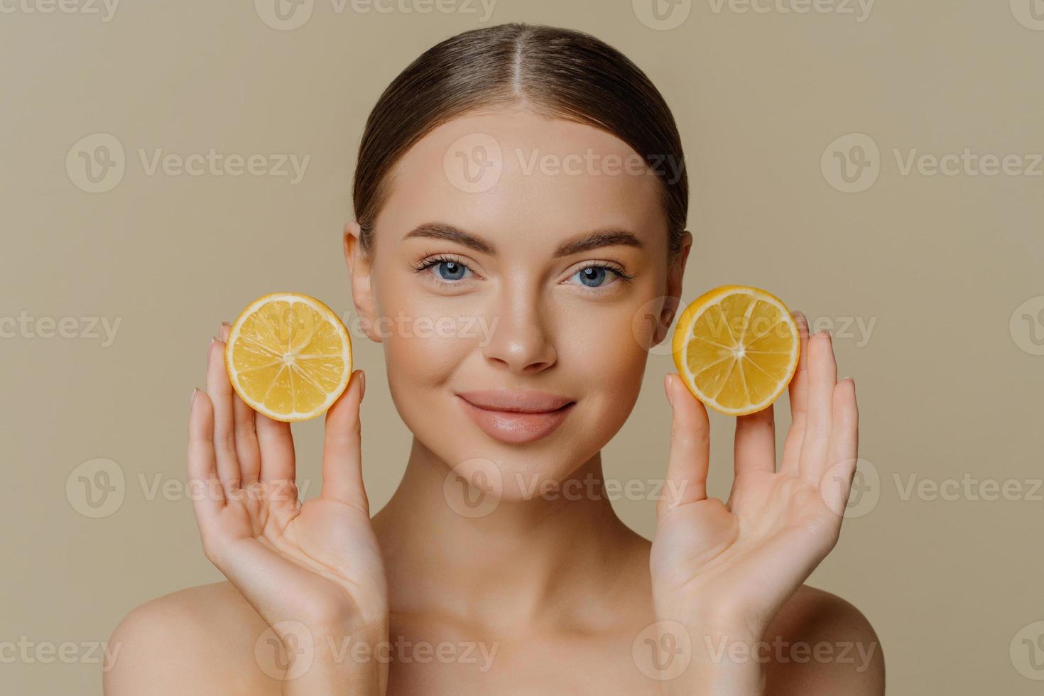 Headshot of beautiful brunette woman holds juicy lemon slices has healthy shiny skin gets vitamins from citrus stands bare shoulders indoor against beige background. Facial treatment concept photo