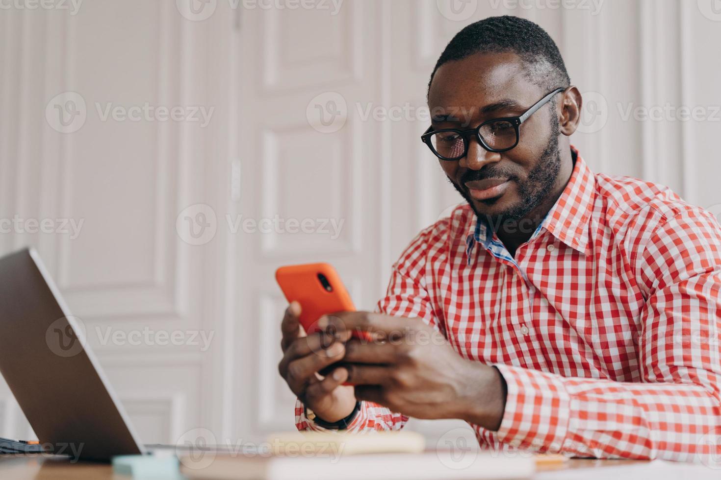 Young handsome african male executive in glasses sitting at office table and using mobile phone photo