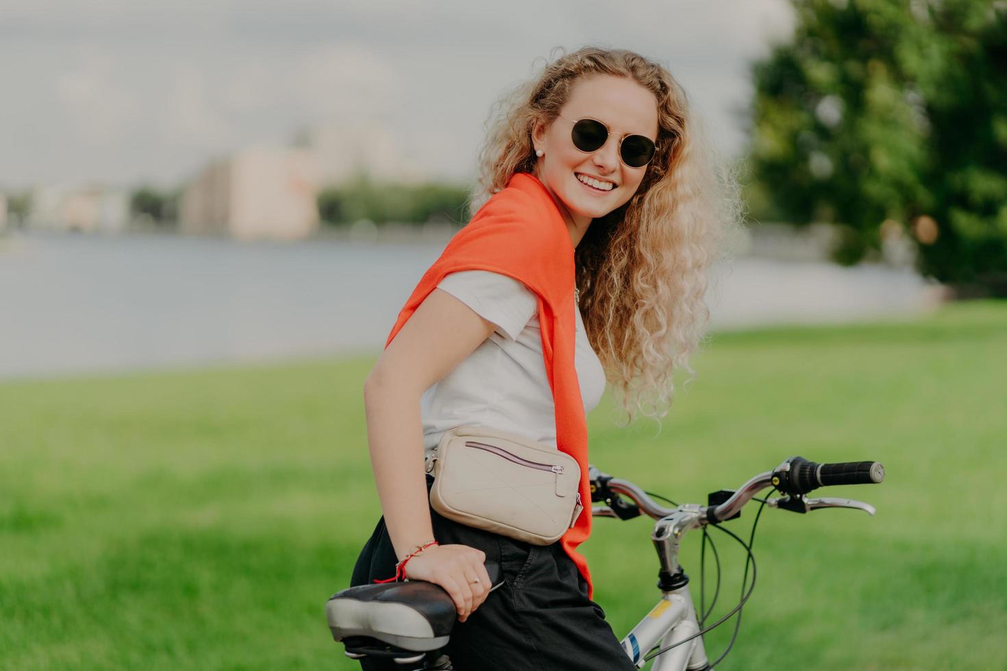 la ciclista satisfecha y rizada disfruta del tiempo libre, viaja en bicicleta, se detiene para descansar un poco, usa gafas de sol, ropa informal de verano, posa entre la hierba y los árboles, fondo verde borroso. foto