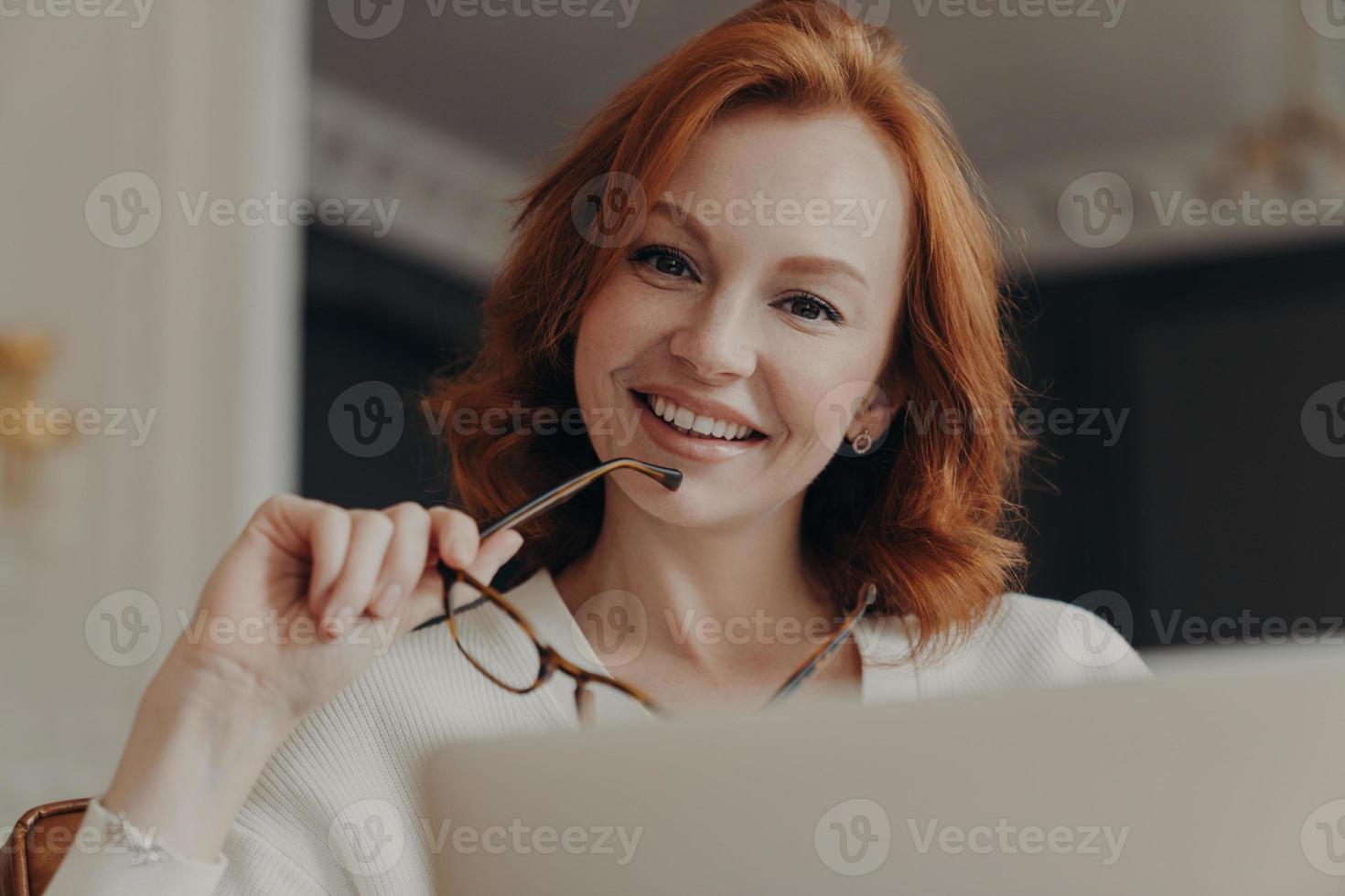 Cheerful redhead female web designer works on freelance, poses at opened laptop computer, holds glasses, shares content in social networks, smiles gently, poses at home. People and technology photo