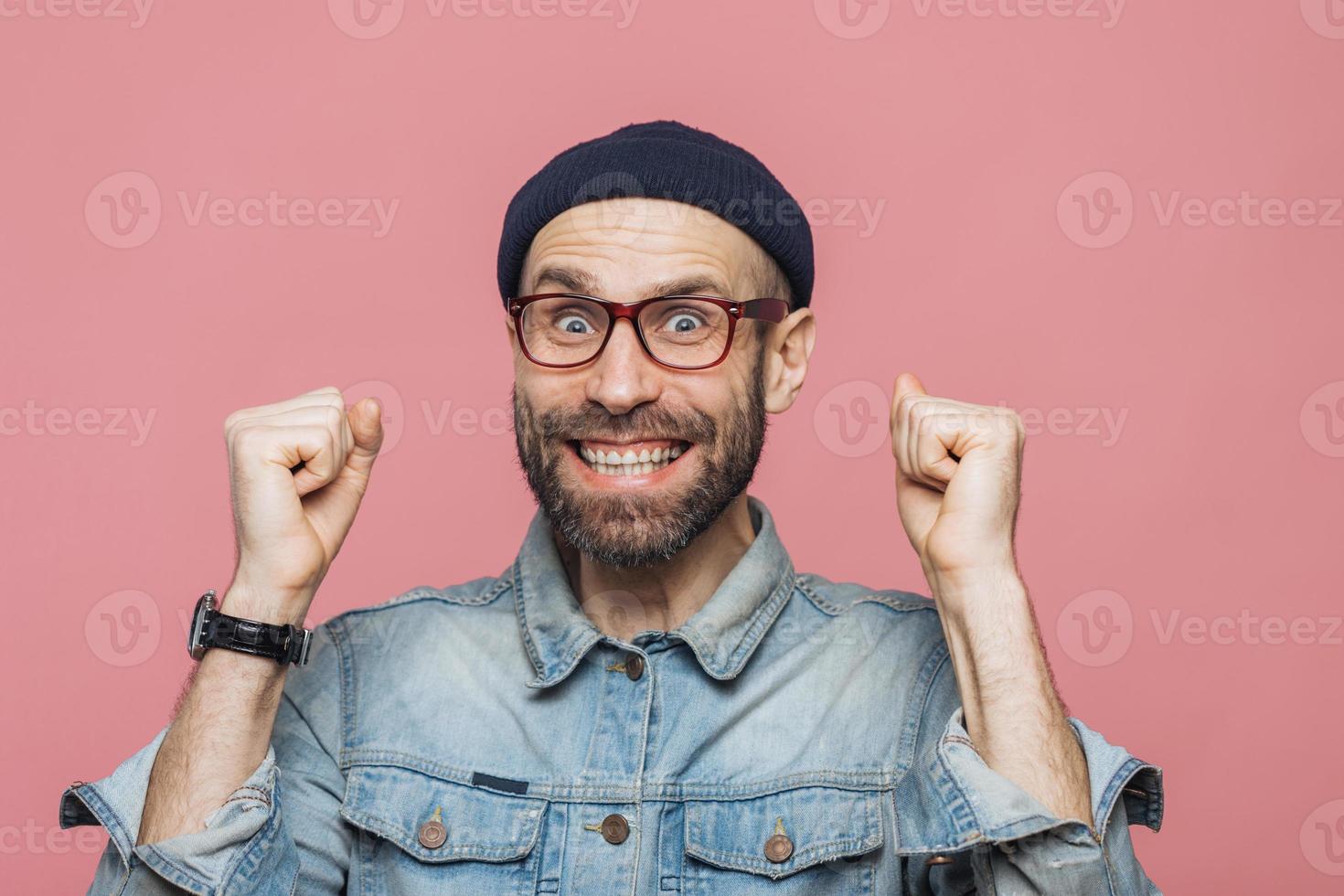 Joyful man with thick beard and mustache clenches fists and looks joyfully at camera, celebrates his victory, has happy expression, isolated over pink background. People, emotions and success photo