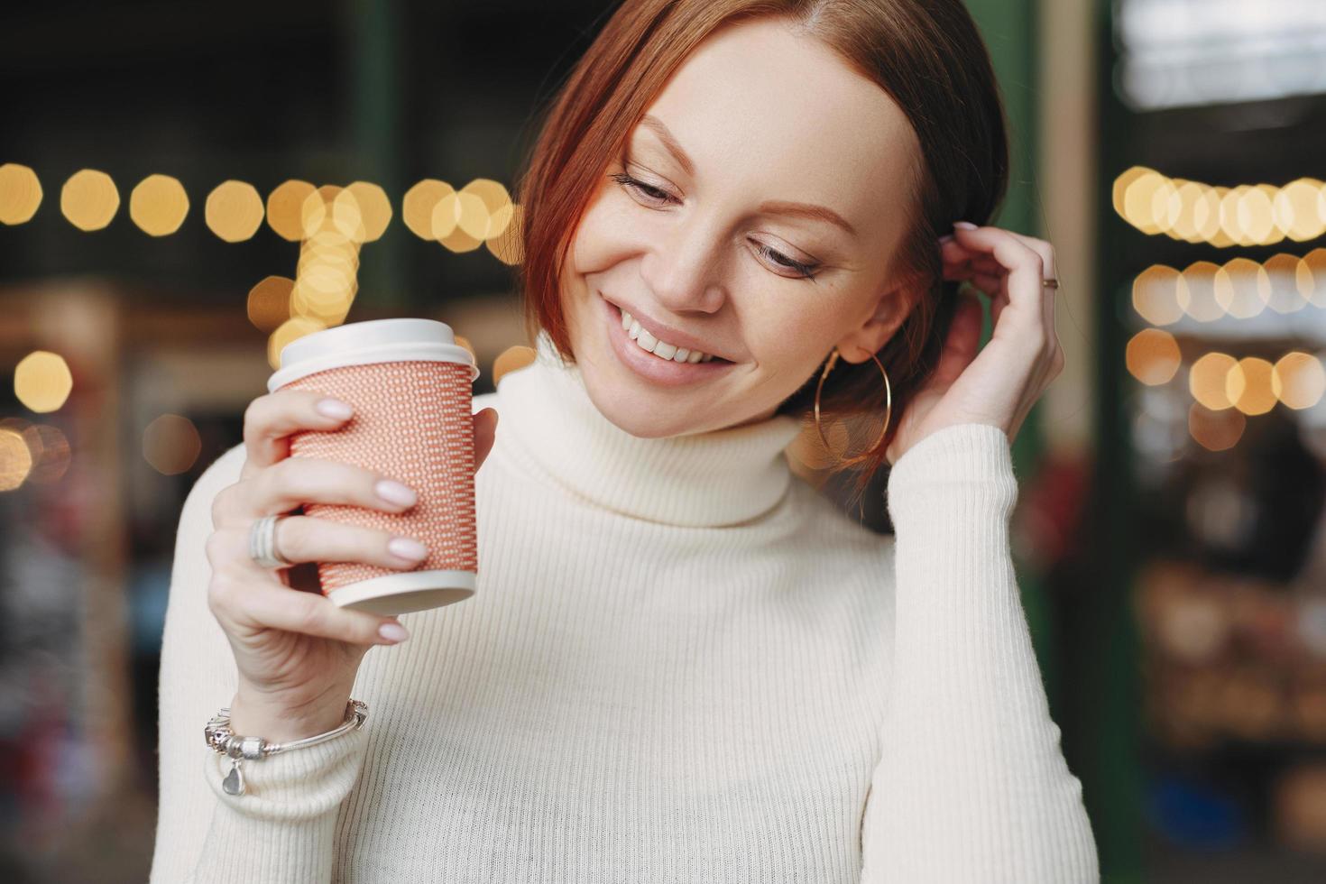 Cropped shot of pleasant looking woman poses in outdoor cafe, holds paper cup of coffee, dressed in white outfit, smiles happily, has talk with colleague or friend during break. Female with beverage photo