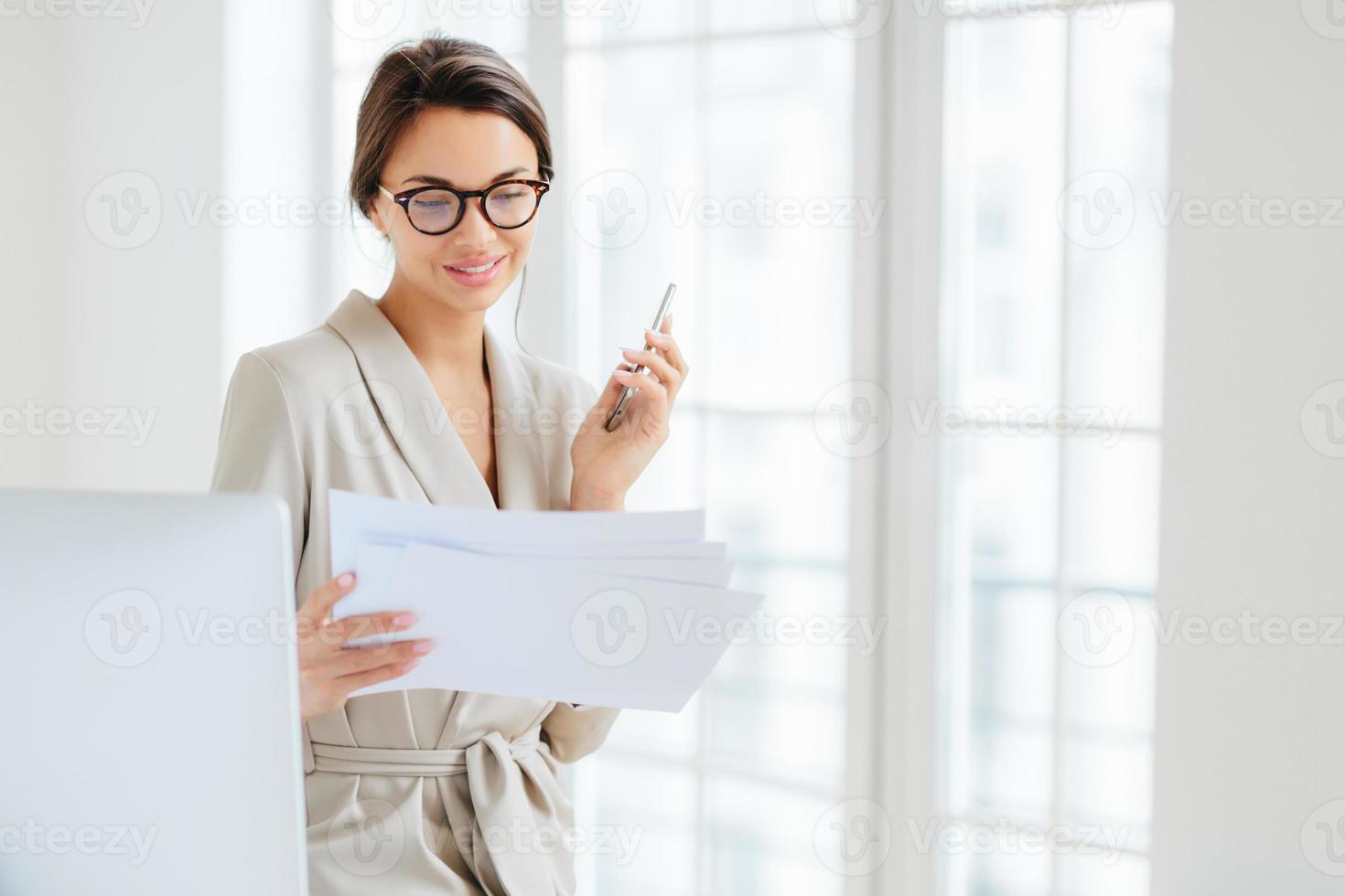 Photo of satisfied young woman with dark hair dressed in business suit, focused in papers, works in office, holds modern cellphone, wears optical glasses for good vision, has pleased expression