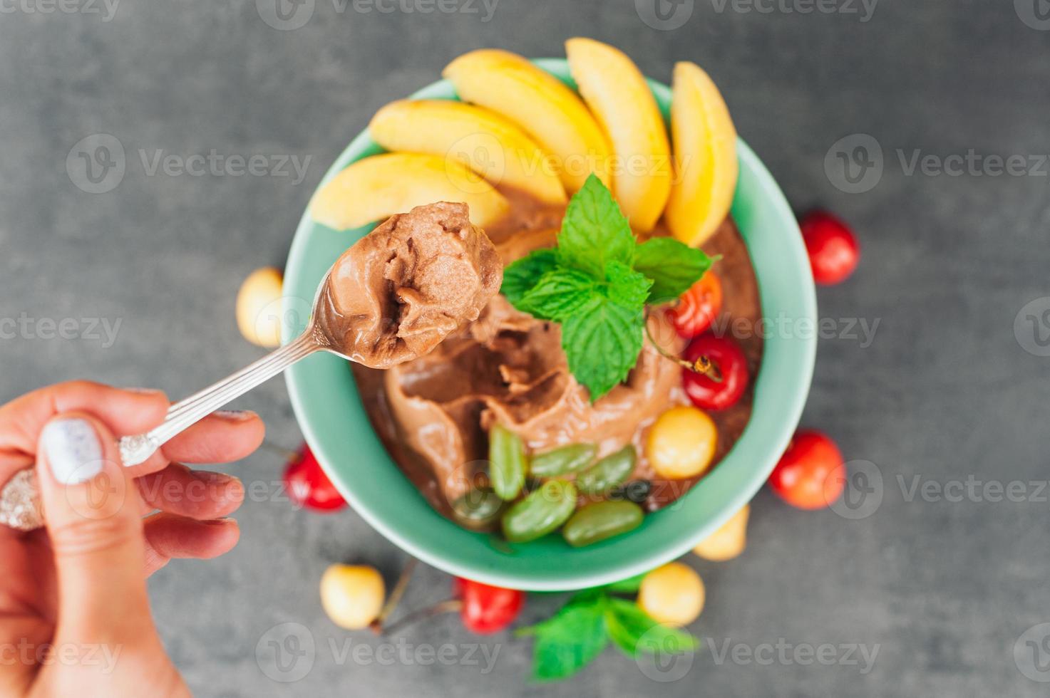 Unknown person holds spoon with chocolate ice cream in hand, bowl with fruits in background. Tasty dessert with banana, grapes, cherry and mint photo