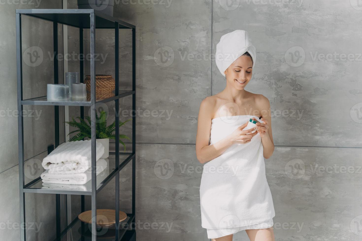 European lady applies cream. Happy young woman wrapped in towel after bathing and hair washing. photo