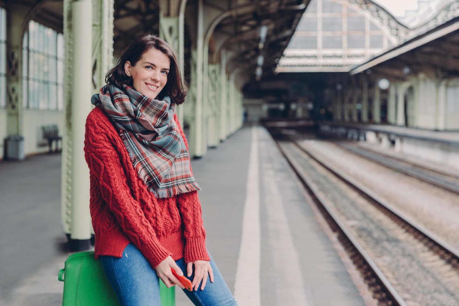 Glad young European woman wears red sweater, scarf, holds mobile phone, sits at bag, enjoy travelling, poses at railway station, smiles happily. People, tourism, travelling and technology concept photo