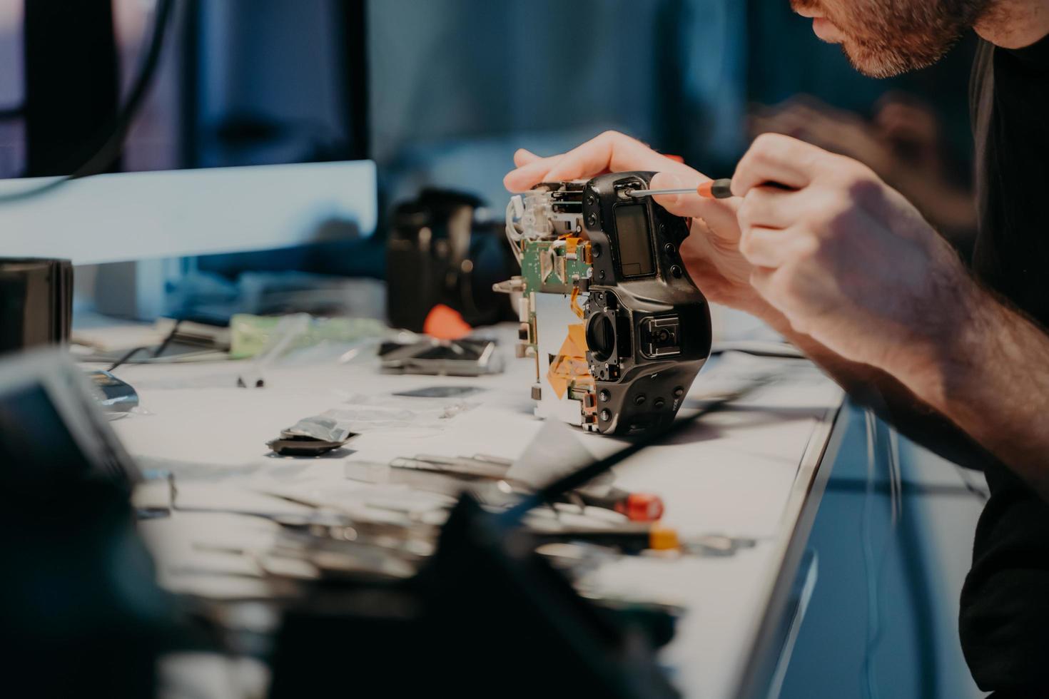 Woking process. Unknown man repairs professional digital camera, uses screwdriver, poses at workplace, holds photocamera disassembled part photo