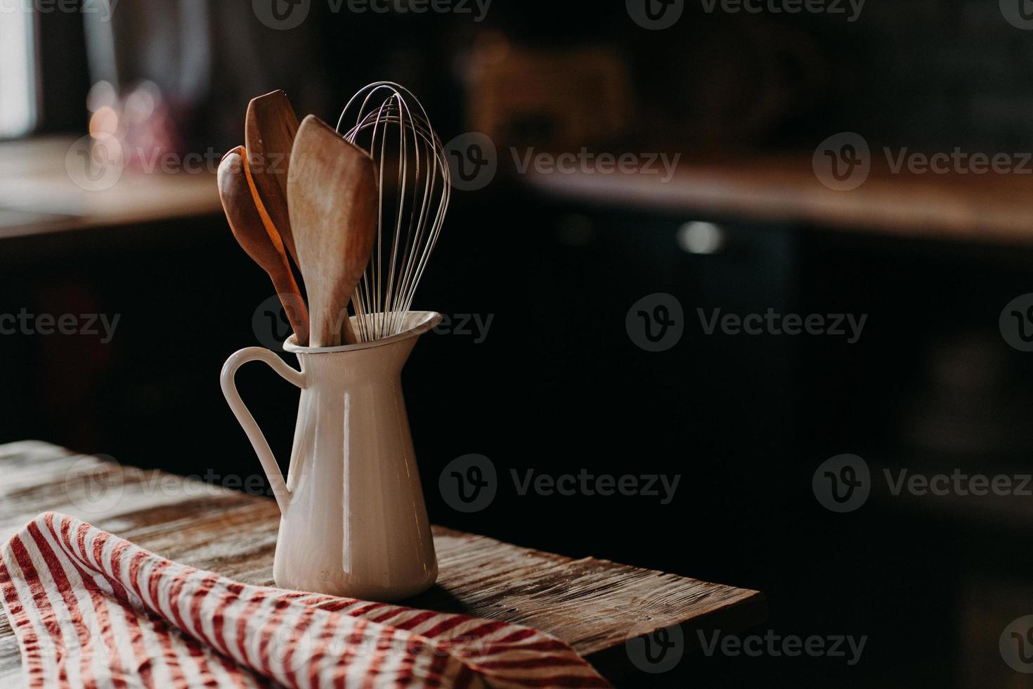 Kitchen accessories on wooden table. Utensils in white ceramic jar against dark background. Rustic style. Dishware for preparing meal. Wooden spoons shpatula and whisk photo