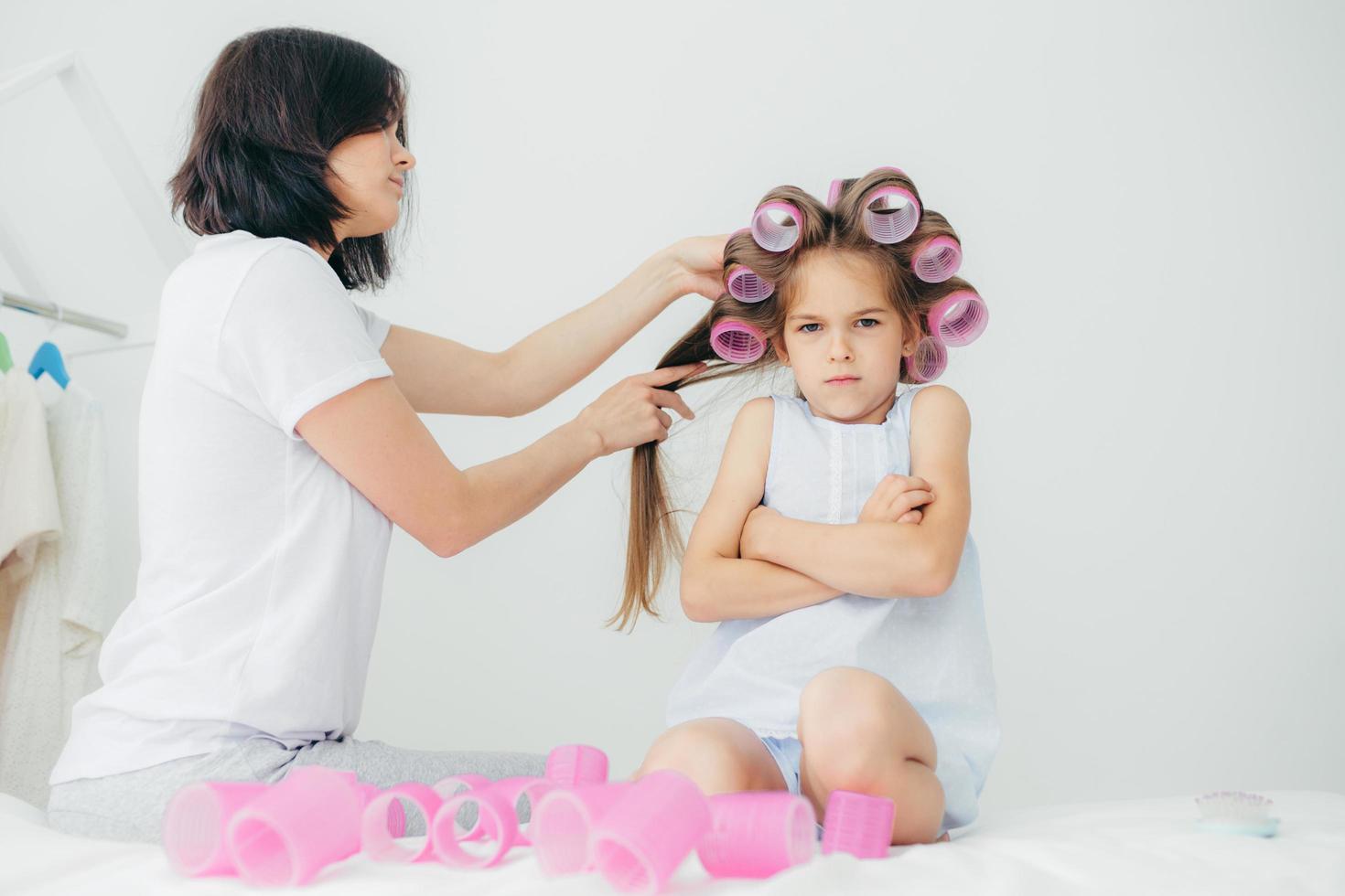 Displeased child keeps hands crossed, looks with sullen expression, being discontent as her mother winds curlers on her hair, doesnt want to have curly hairstyle, pose against white background photo