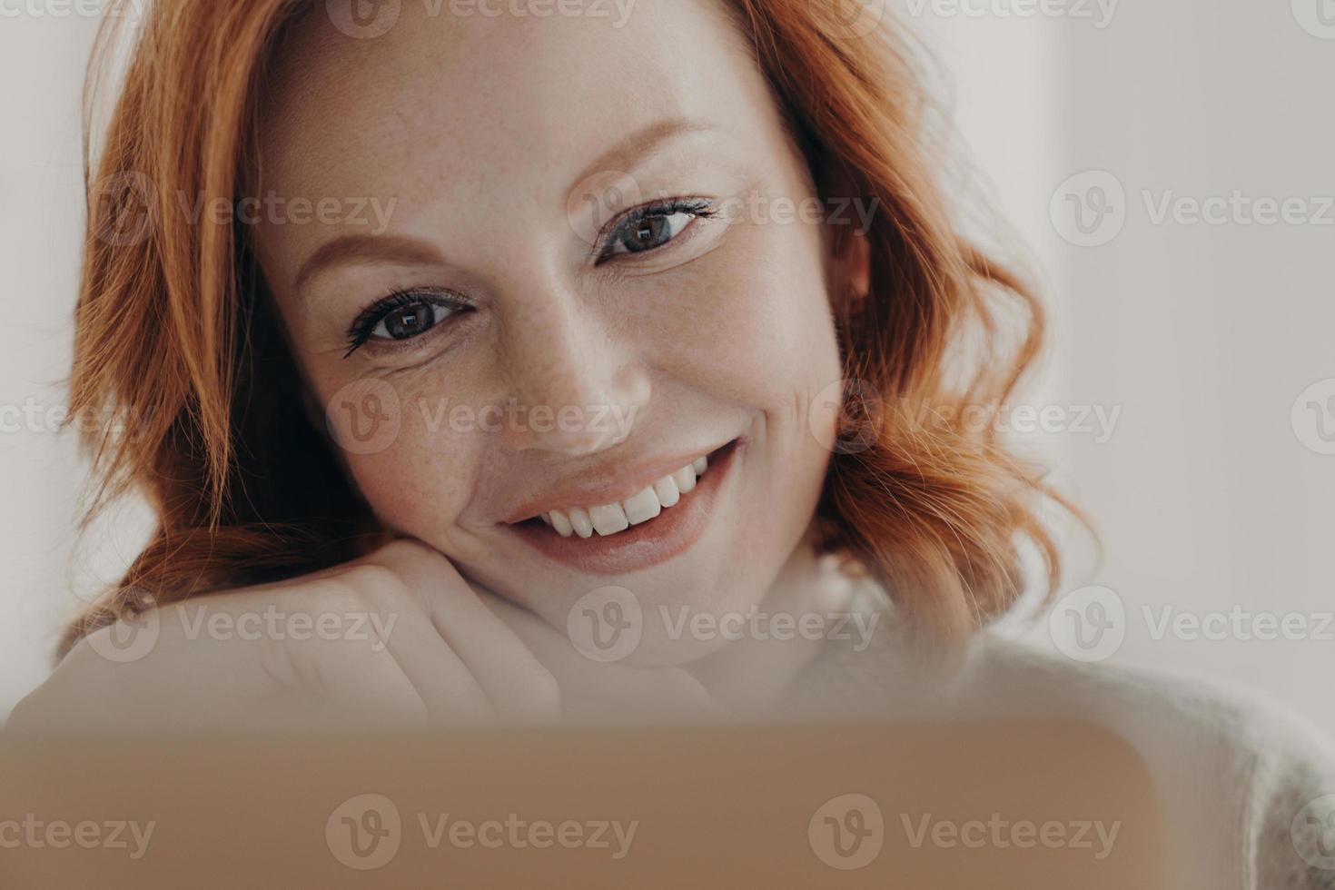 Close up shot of positive redhead woman with freckled skin and toothy smile, concentrated at display of laptop computer, satisfied with online business, checks received message, has remote job photo