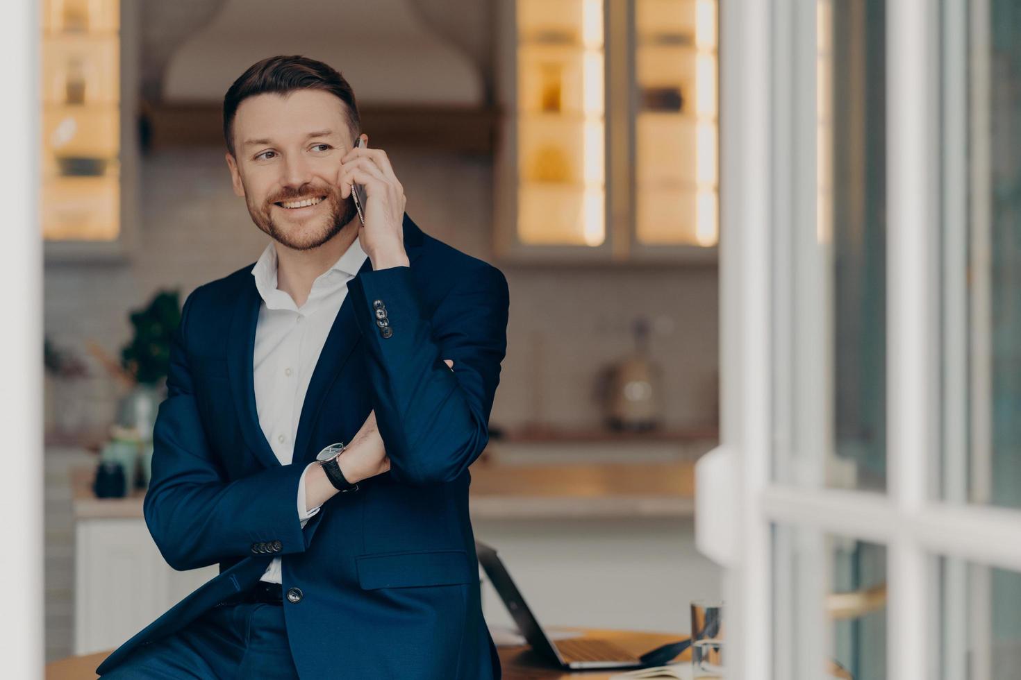 Corporate communication. Smiling professional bearded male entrepreneur has succssful business conversation keeps modern smartphone near ear poses near desktop dressed in formal suit focused away. photo