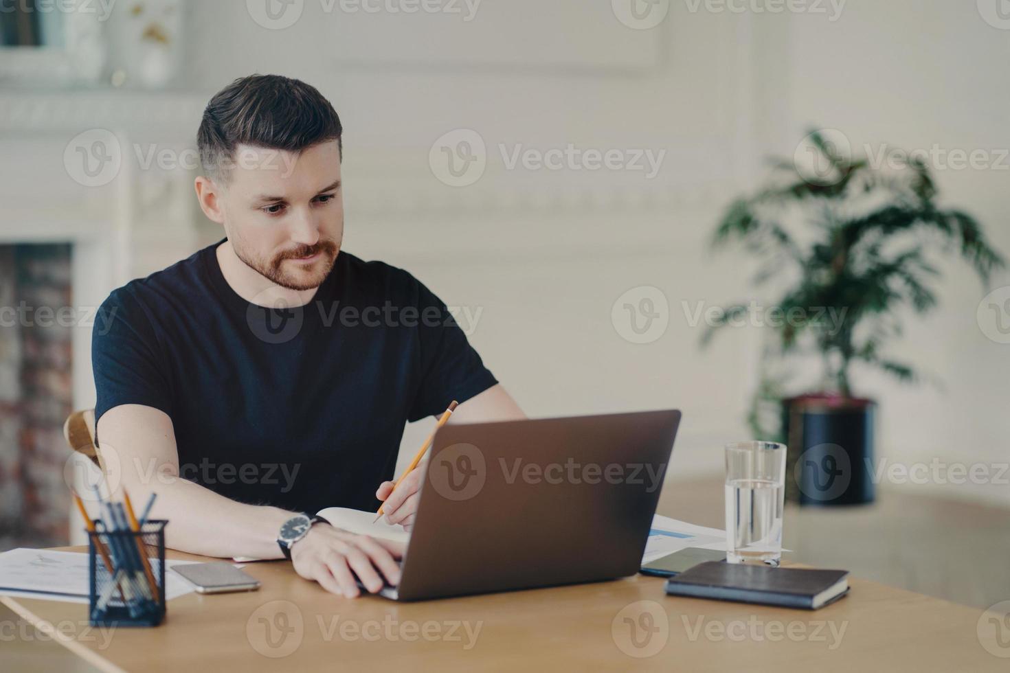 Serious bearded man writes informative notes while watching webinar on laptop computer works remotely dressed in casual t shirt poses at workplace against home office interior. Modern workspace photo