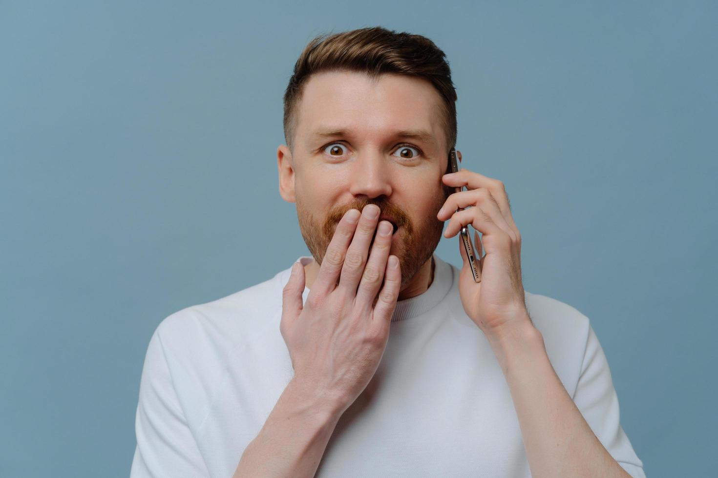 Astonished surprised bearded adult man covers mouth looks with amazement at camera has telephone conversation keeps smartphone near ear isolated over blue background. Human reactions concept photo