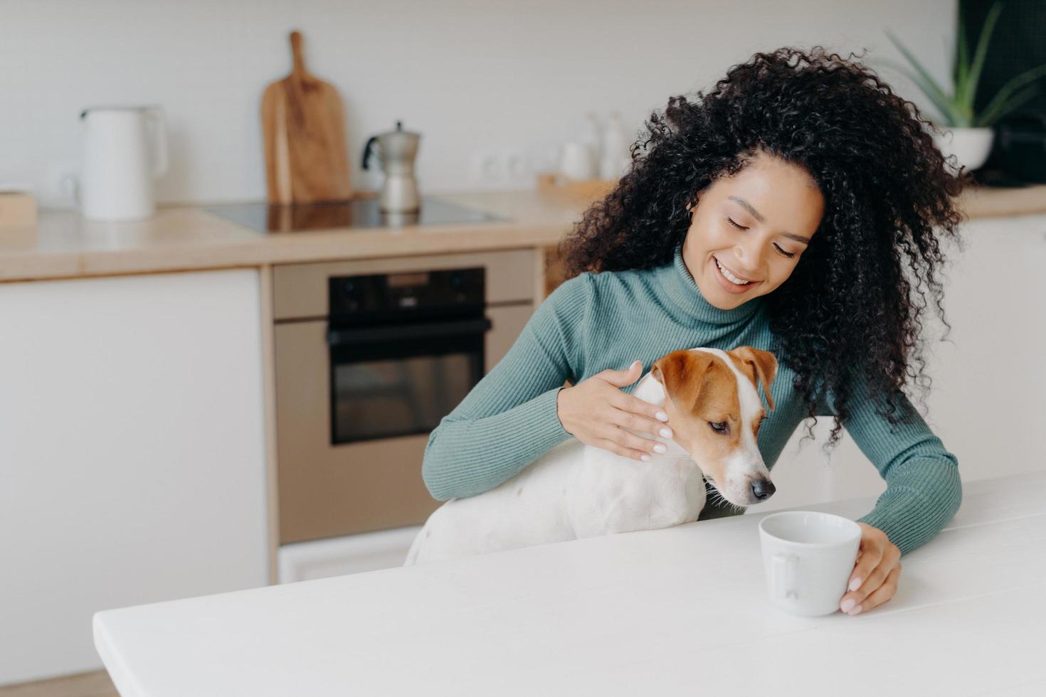 Happy Afro African woman with curly hairstyle treats dog in kitchen, pose at white table with mug of drink, enjoy domestic atmosphere, have breakfast together. People, animals, home concept. photo