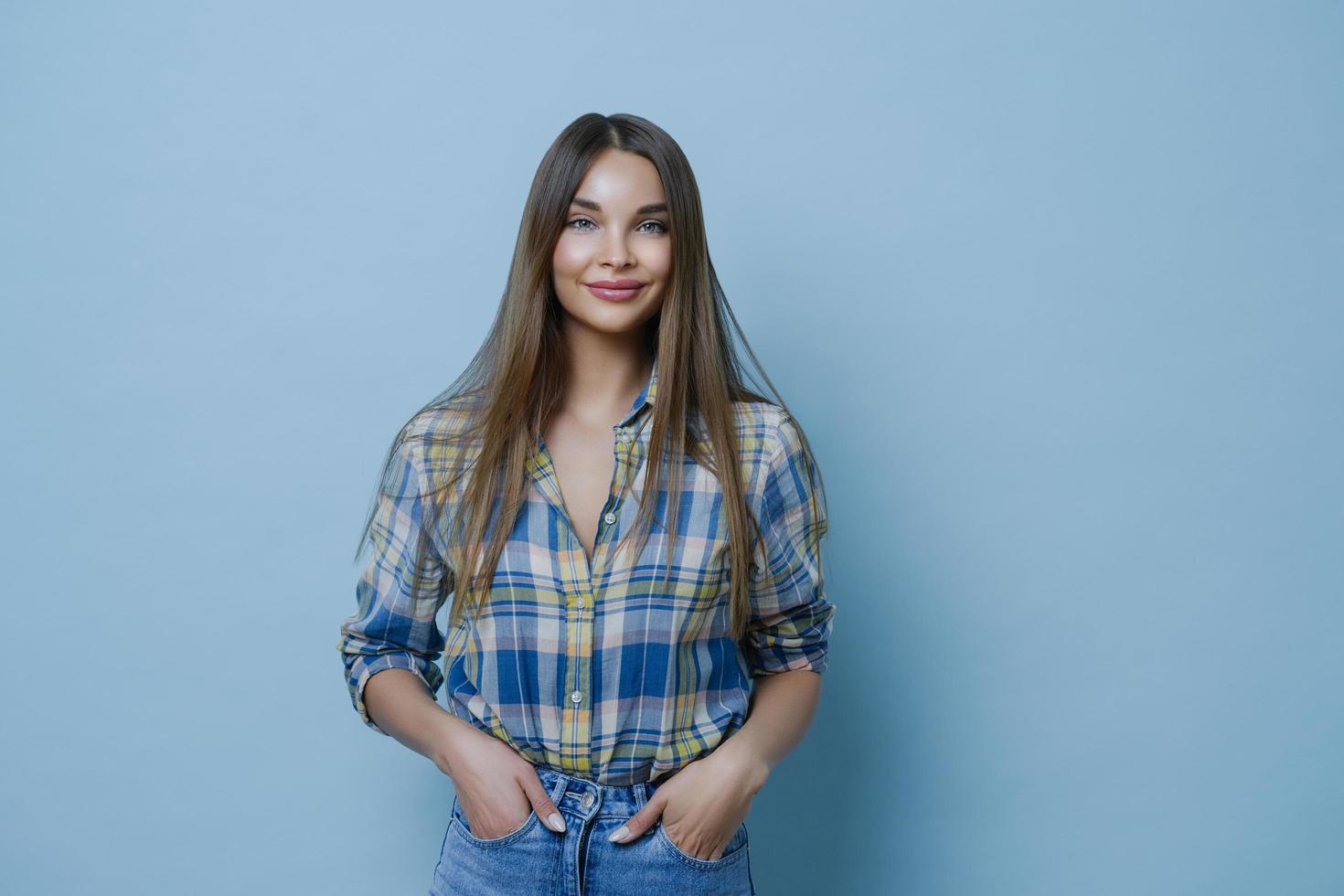 Attractive student glad to talk about part time job, keeps hands in pockets of jeans, being happy because of positive news, has pleasant smile, isolated over blue studio background. People and style photo