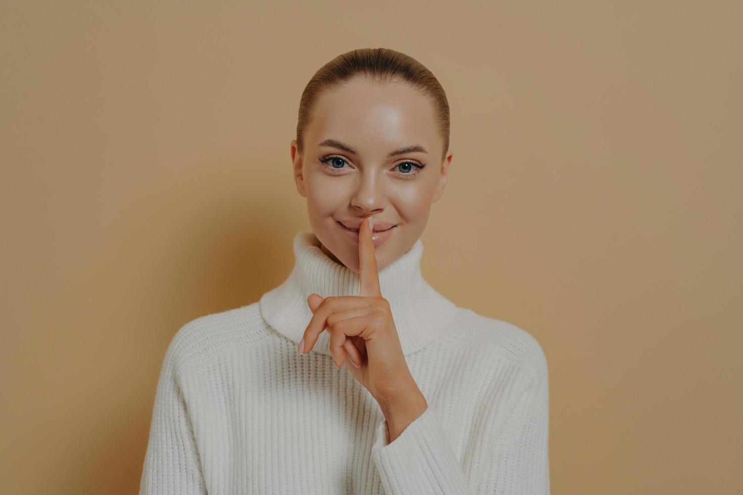 Keep it secret. Young attractive smiling lady saying be quiet with finger on lips, isolated on beige photo