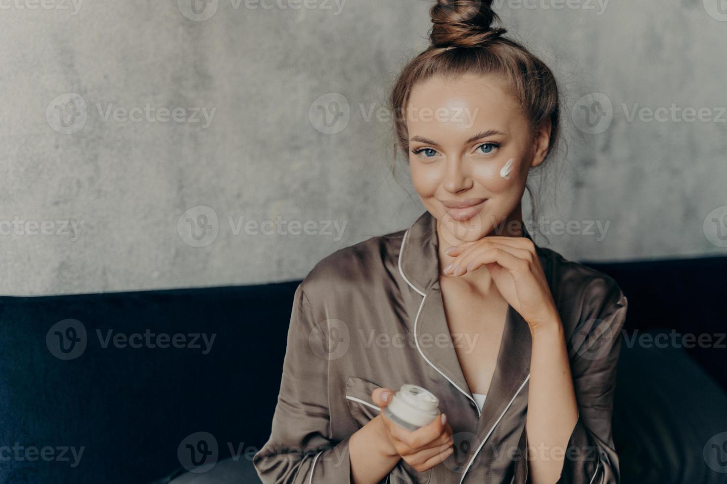 Attractive young female model in brown silk pajama sitting on bed with face cream photo