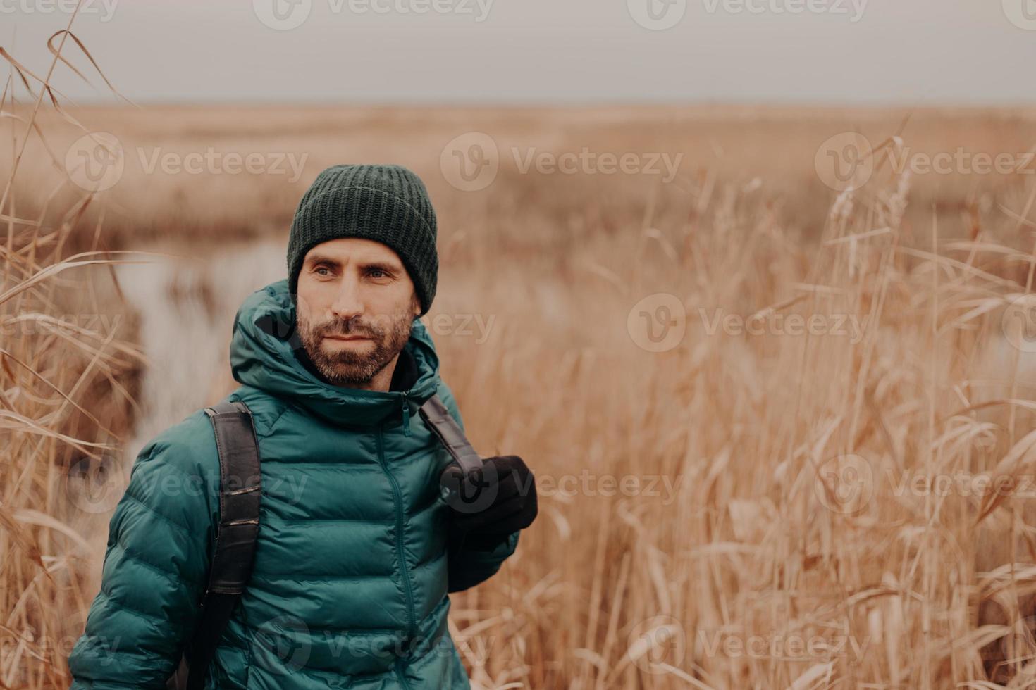 Horizontal shot of contemplative good looking man has stubble, wears hat, jacket and gloves, stands near wheat field background with free space on right side. People, leisure and walk concept photo