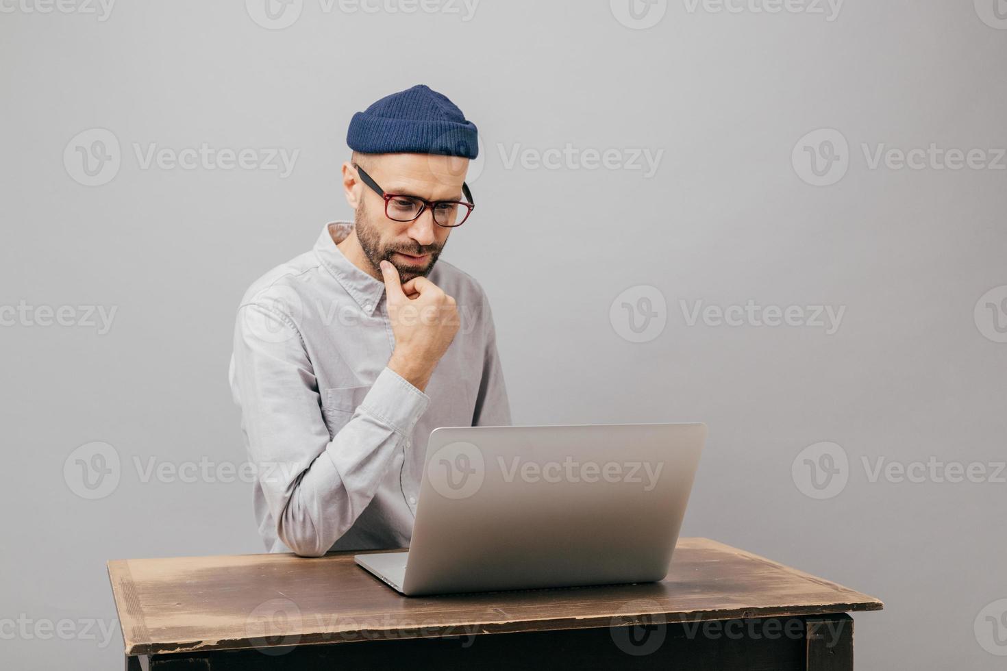 Image of unshaven male owner of trading company searches information about marketing agency, thinks about formal meeting, wears glasses and elegant clothes, sits at old desk over grey background photo
