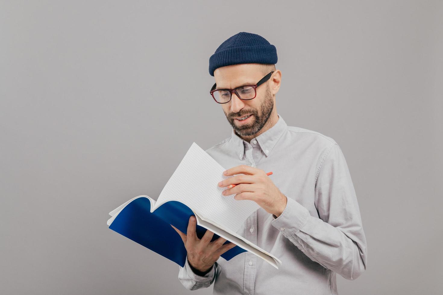 Photo of handsome unshaven young guy holds notebook, turns over page, writes records and planning for next week, wears optical glasses and white formal shirt, models over grey background alone