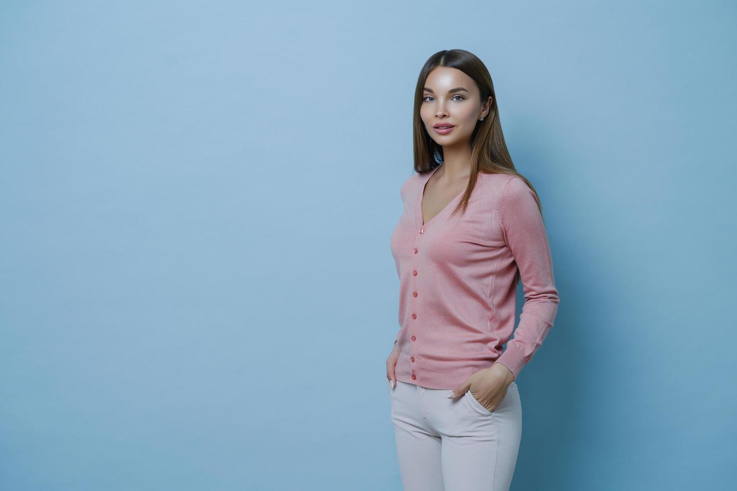 Sideways shot of beautiful dark haired young European woman wears stylish jumper, keeps hands in pocket of white trousers, poses against blue background, blank space for your advertising content. photo