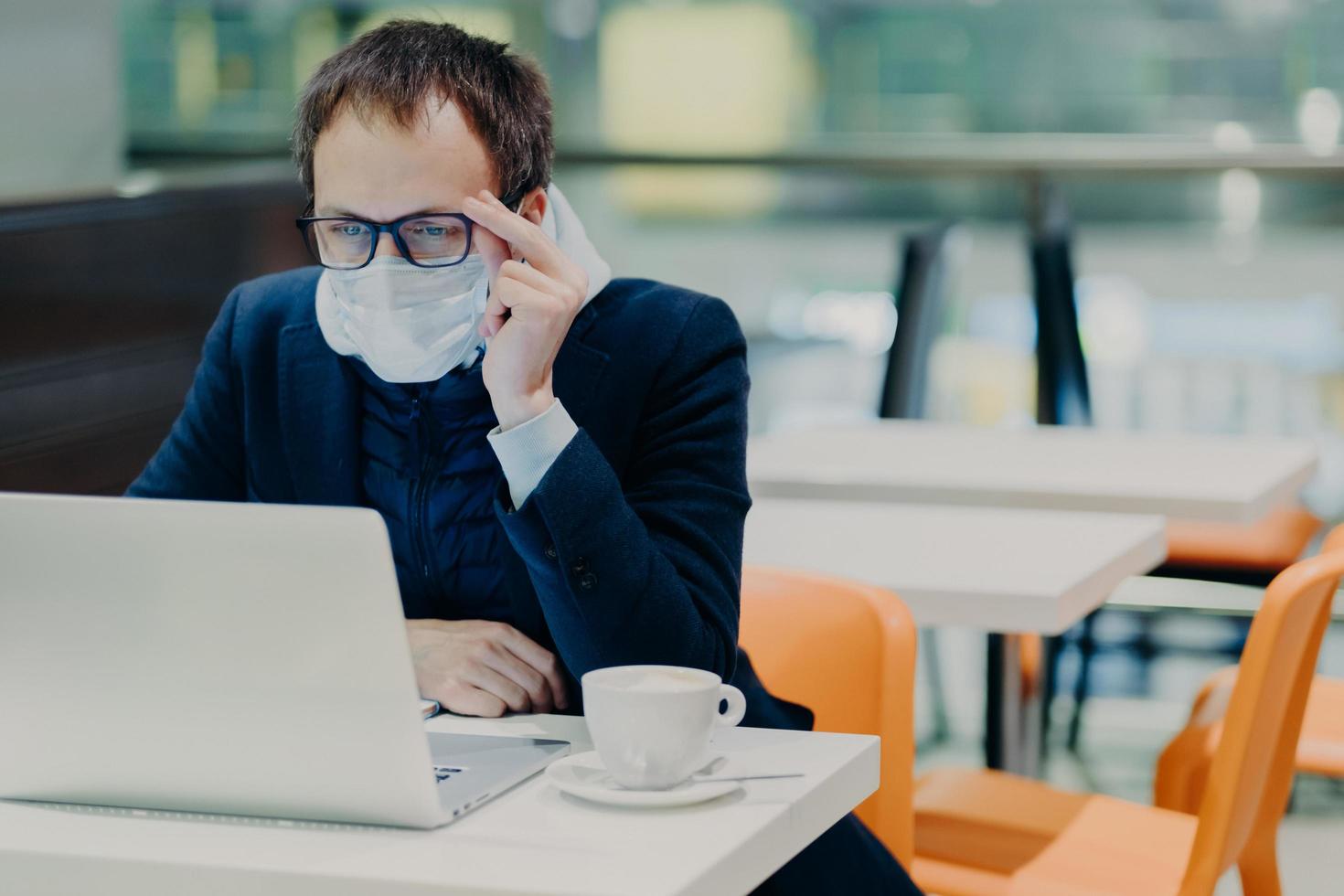 Man in medical mask and spectacles, sits in front of modern laptop computer, poses in cozy cafeteria, reads news about coronavirus, concerned about health, drinks coffee. Epidemic quarantine photo