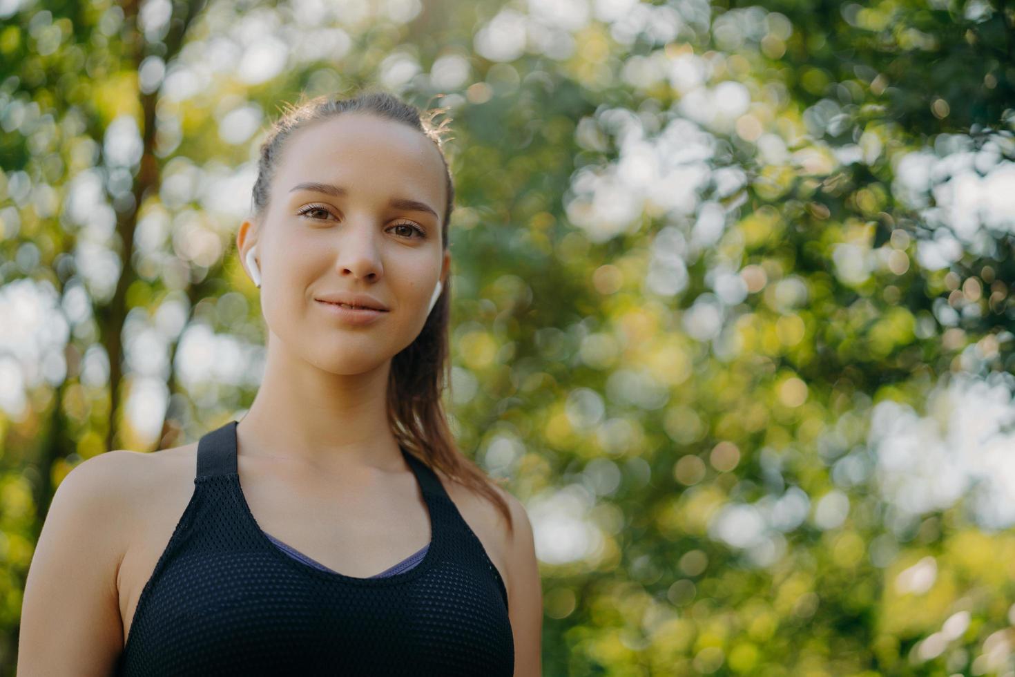 Horizontal shot of beautiful young woman with dark combed hair enjoys outdoor workout listens music in wireless earphones poses against green blurred nature background. Sport fitness healthy lifestyle photo