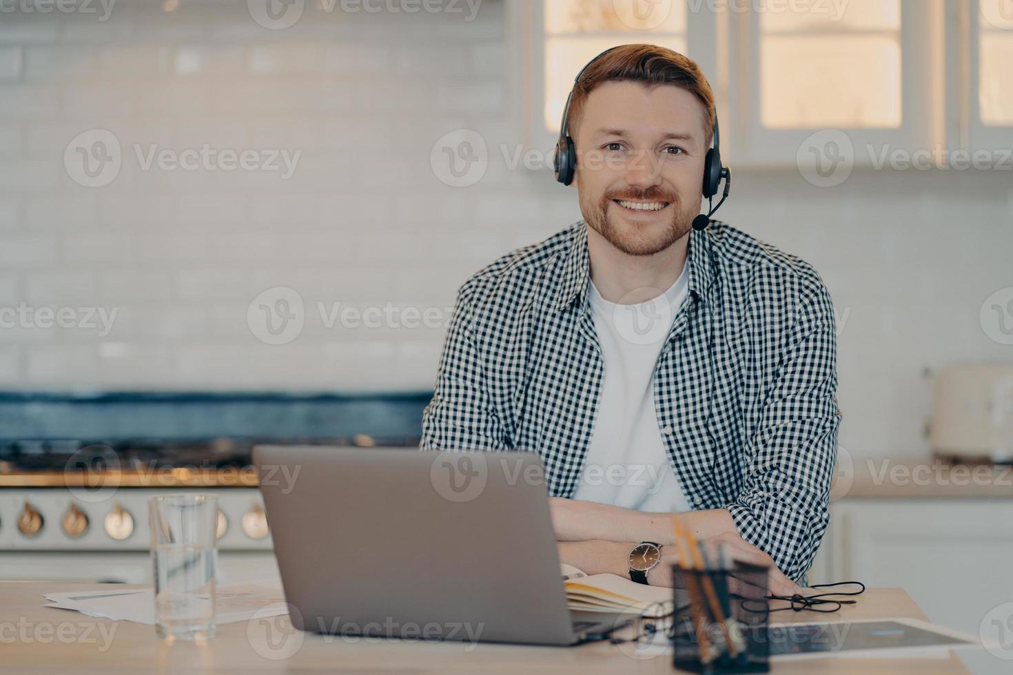 Handsome glad man customer support manager gives advice or consultation to clients works online from home poses at home kitchen wears wireless headphones uses modern laptop computer. Remote job photo