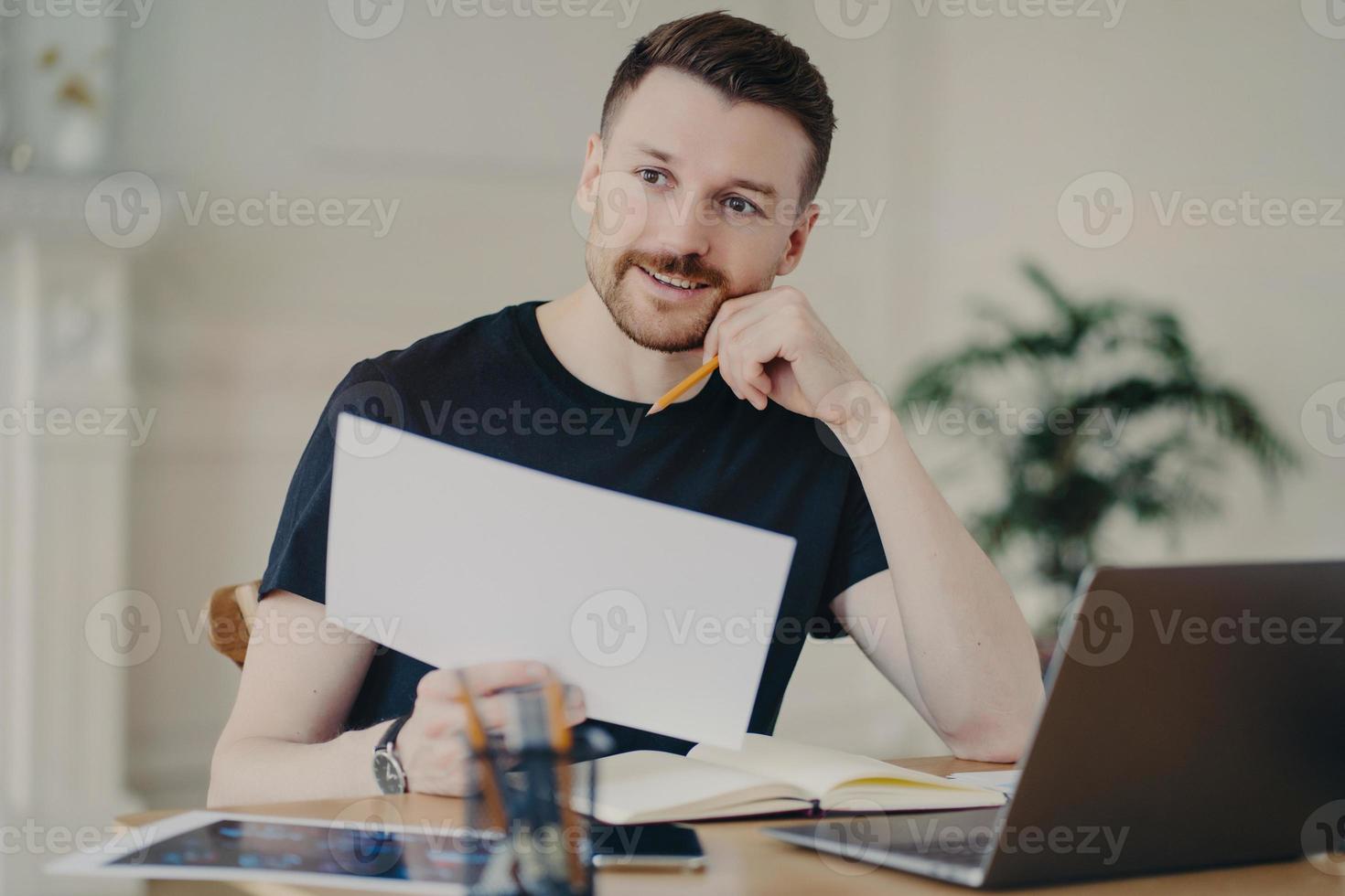 Glad handsome European businessman holds paper document poses in home interior at desktop surrounded by modern technologies checks information has remote work looks thoughtfully into distance photo