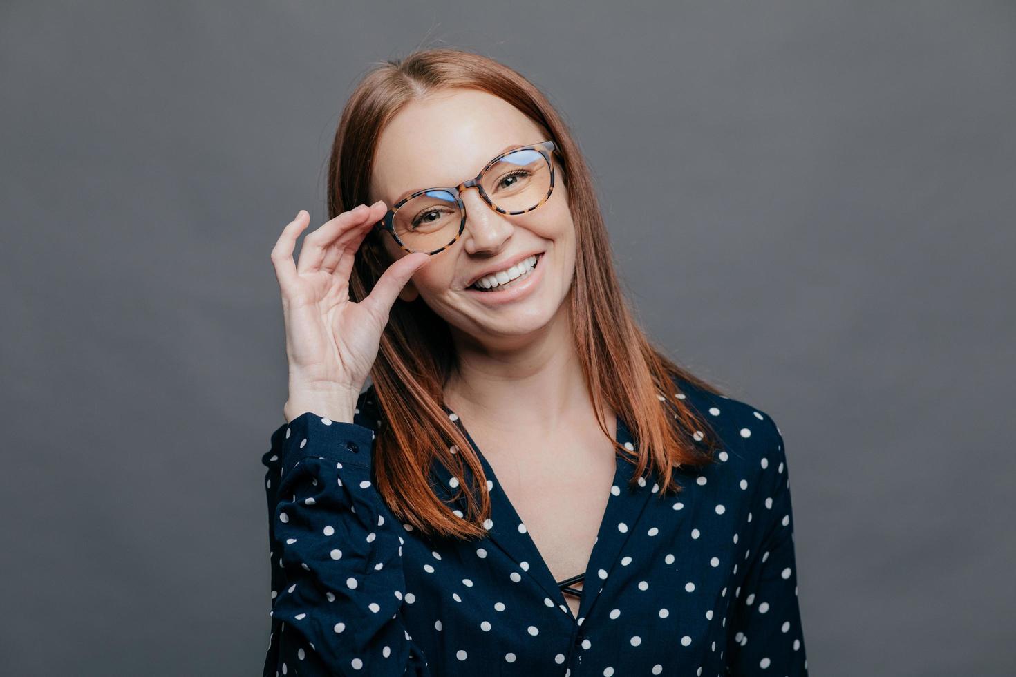 mujer sonriente con expresión facial complacida, mantiene la mano en el borde de los espectáculos, usa blusa de lunares negros, se regocija de la promoción en el trabajo, posa sobre fondo gris. gente, concepto de felicidad foto