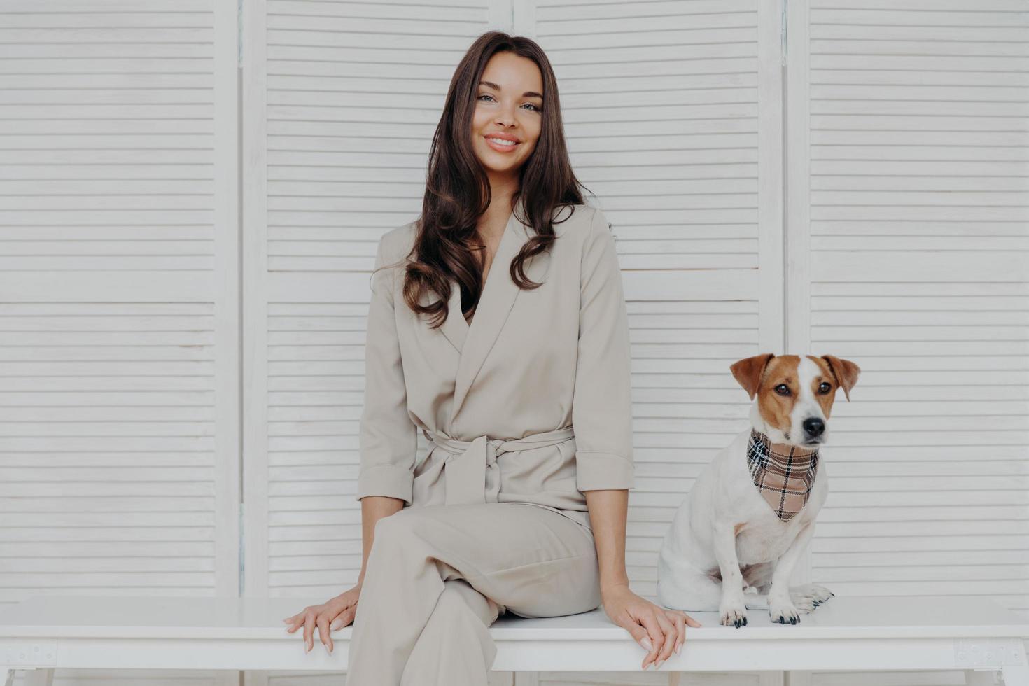 Image of attractive brunette European woman sits together with pedigree dog, dressed in fashionable clothes, looks with gentle smile at camera. People, animal care and responsibility concept photo