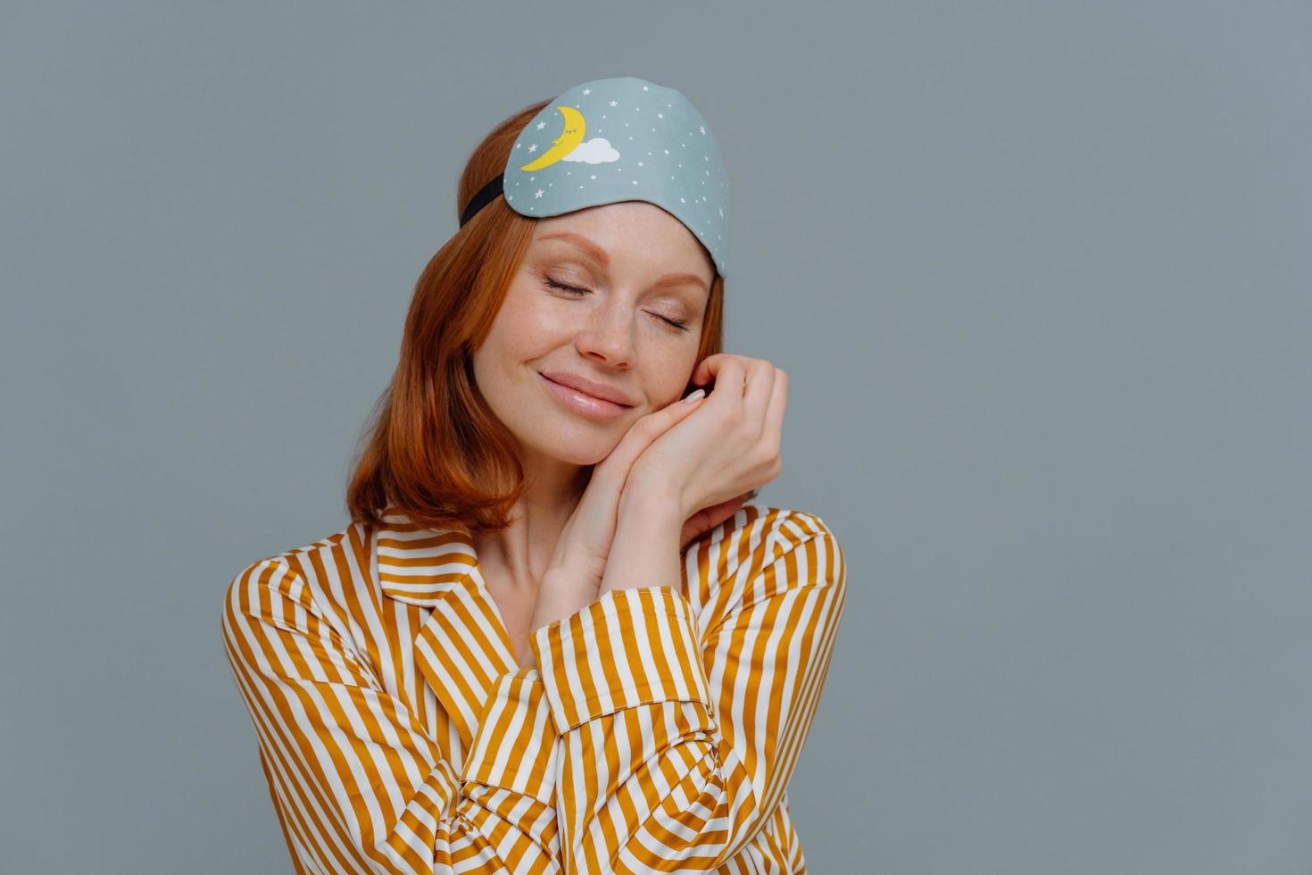 Pretty foxy freckled woman leans at palms, keeps eyes closed, wears sleepmask and stiped pajamas, dreams about something pleasant, stands indoor over grey wall, with copy space for promotion photo