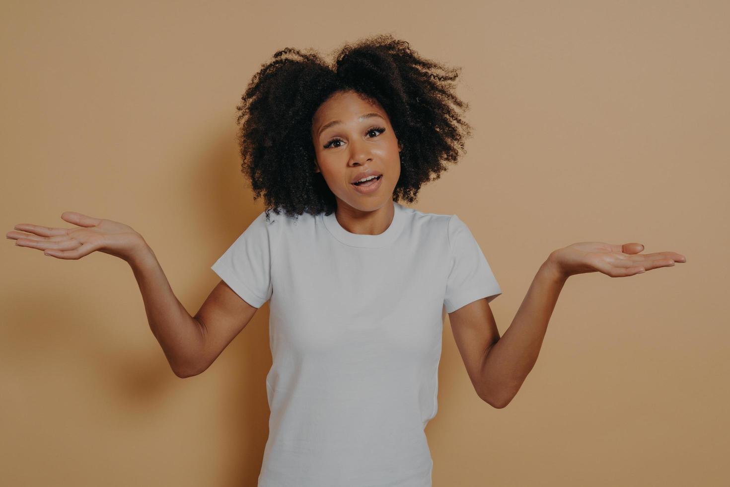 Unsure african woman woman shrugging shoulders and spreading arms sideways, isolated over beige wall photo