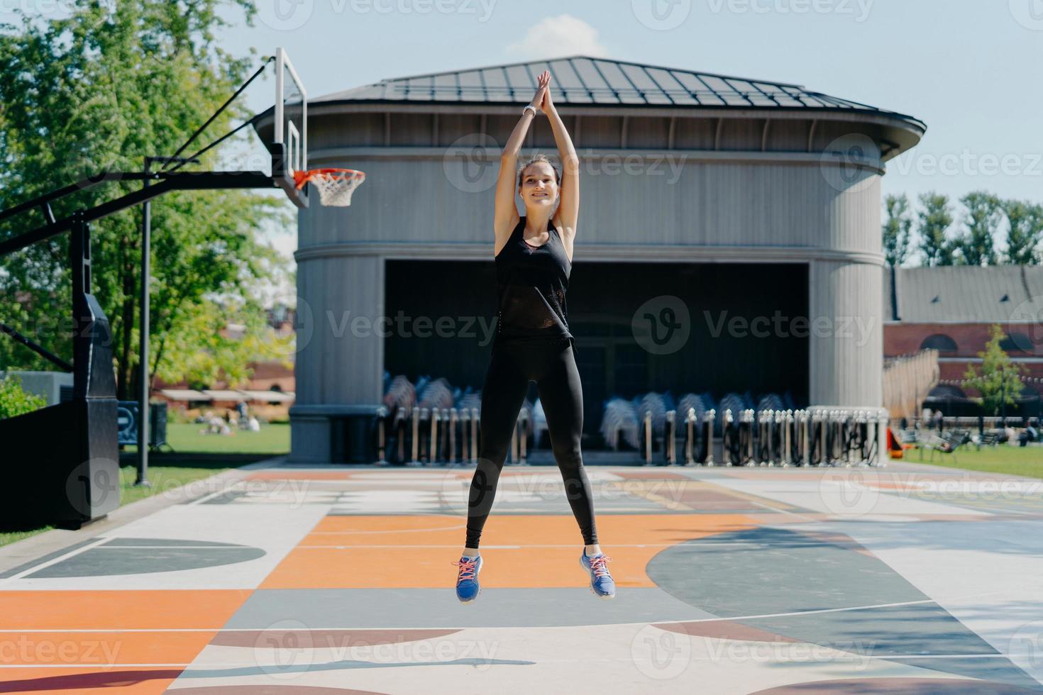 Athletic young woman in black activewear jumps high keeps arms raised up clasps hands warms up before jogging has training on stadium outdoors has cardio burns fat and calories for weight loss photo