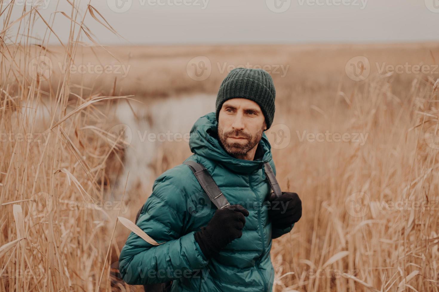 Shot of handsome young man with dark stubble, carries backpack, looks back, notices something, has thoughtful expression, spends free time in open air. Autumn time. People and adventure concept photo