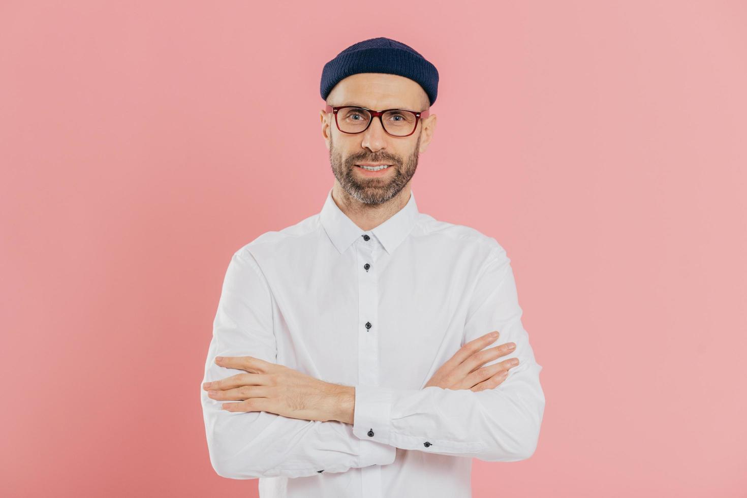Indoor shot of self confident unshaven man looks happily at camera, keeps arms folded, wears headgear and shirt, listens attentively interlocutor, isolated over pink background. Masculinity concept photo