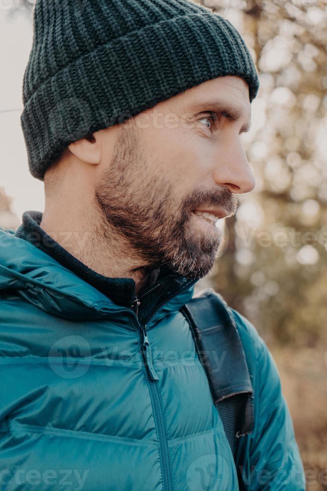 Sideways shot of unshaven man with dark thick beard, wears warm har and anorak, looks pensively aside, poses outside, dreams about something has outdoor stroll in unknown place alone, active lifestyle photo