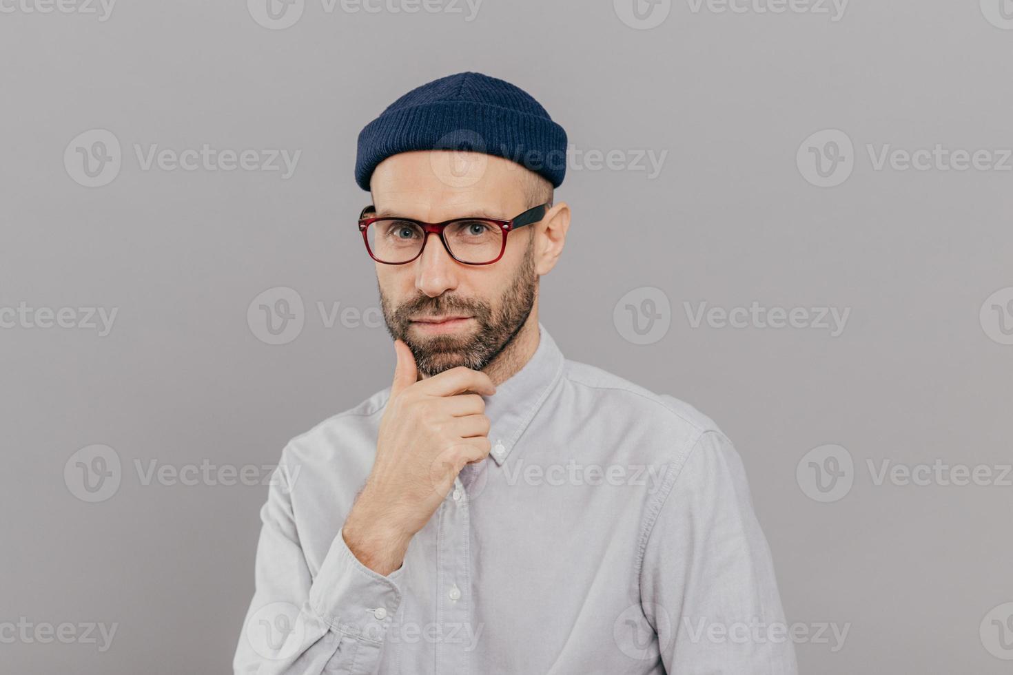 Blue eyed male adult holds chin and looks attentively at camera, ponders on way out or solution, wears spectacles, headgear and formal shirt, models over grey studio wall. Businessman poses indoor photo