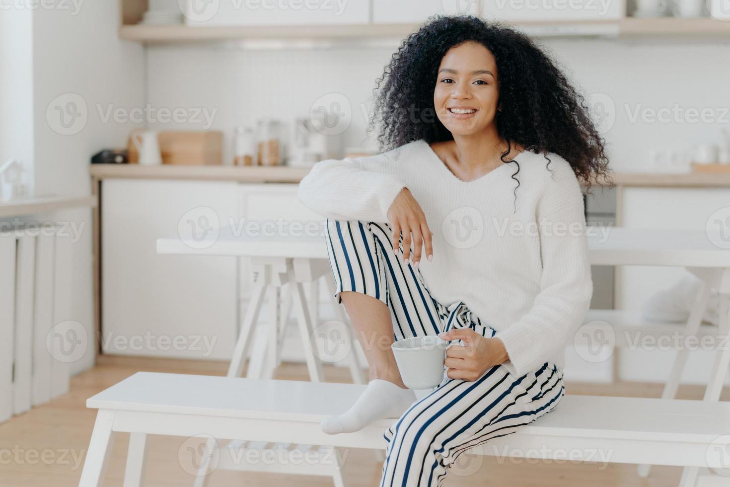 Glad African American woman holds cup of beverage, leans at knee, stylish jumper and striped trousers, smiles pleasantly, spends leisure time at home, sits at bench in kitchen 8836419