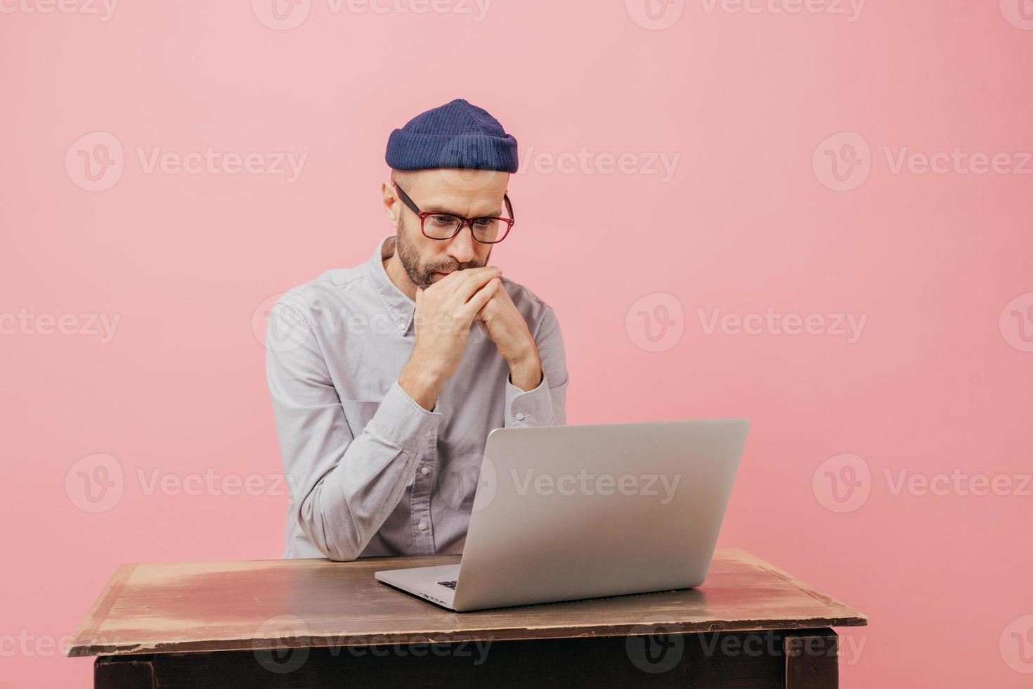 Horizontal shot of concentrated male worker reads statistics attentively, focused in laptop computer, learns something online, wears spectacles, hat and white shirt, isolated over pink wall. photo