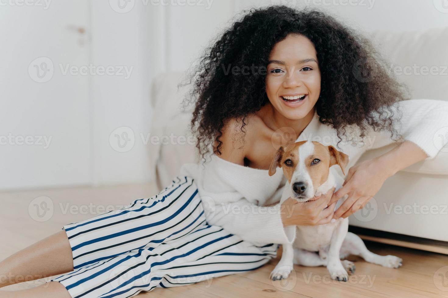 Image of happy curly woman plays with funny little dog, pose on floor in spacious room, sofa near, smiles broadly, embraces pet with love, dressed in fashionable clothes, has fun with animal photo