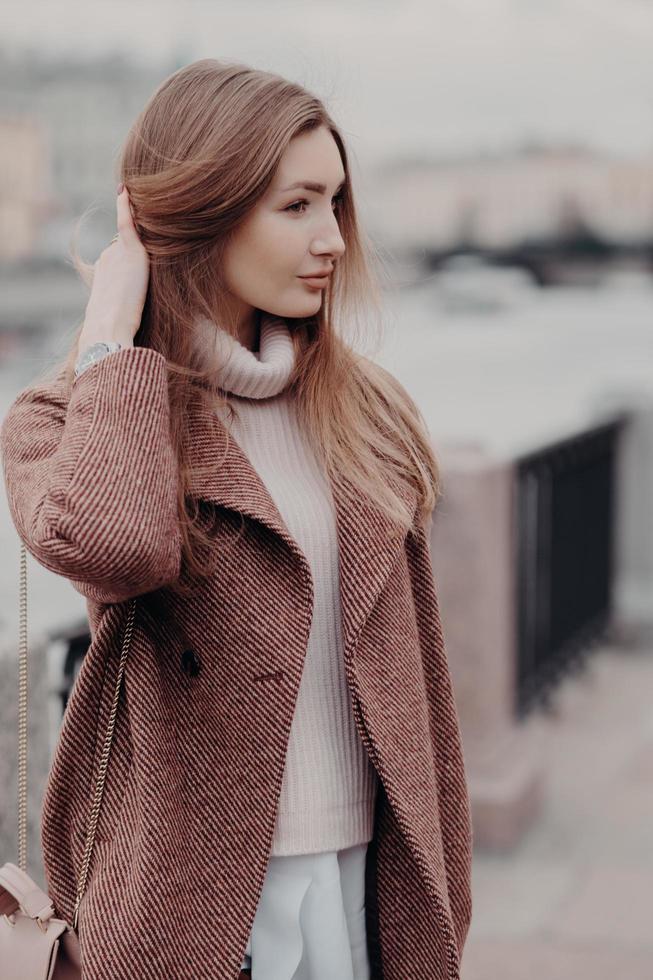 Vertical shot of fashionable Caucasain woman looks aside, wears fashionable warm coat, poses outdoor, enjoys day off, has date. Beautifu young lady goes to work, carries womans bag. Urban style photo