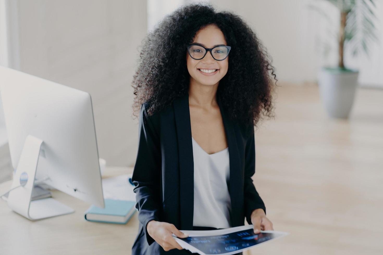 Pleased skilled dark skinned curly woman holds some papers, poses in modern office interior, wears transparent glasses and formal suit, stands near table with computer, studies documentation photo