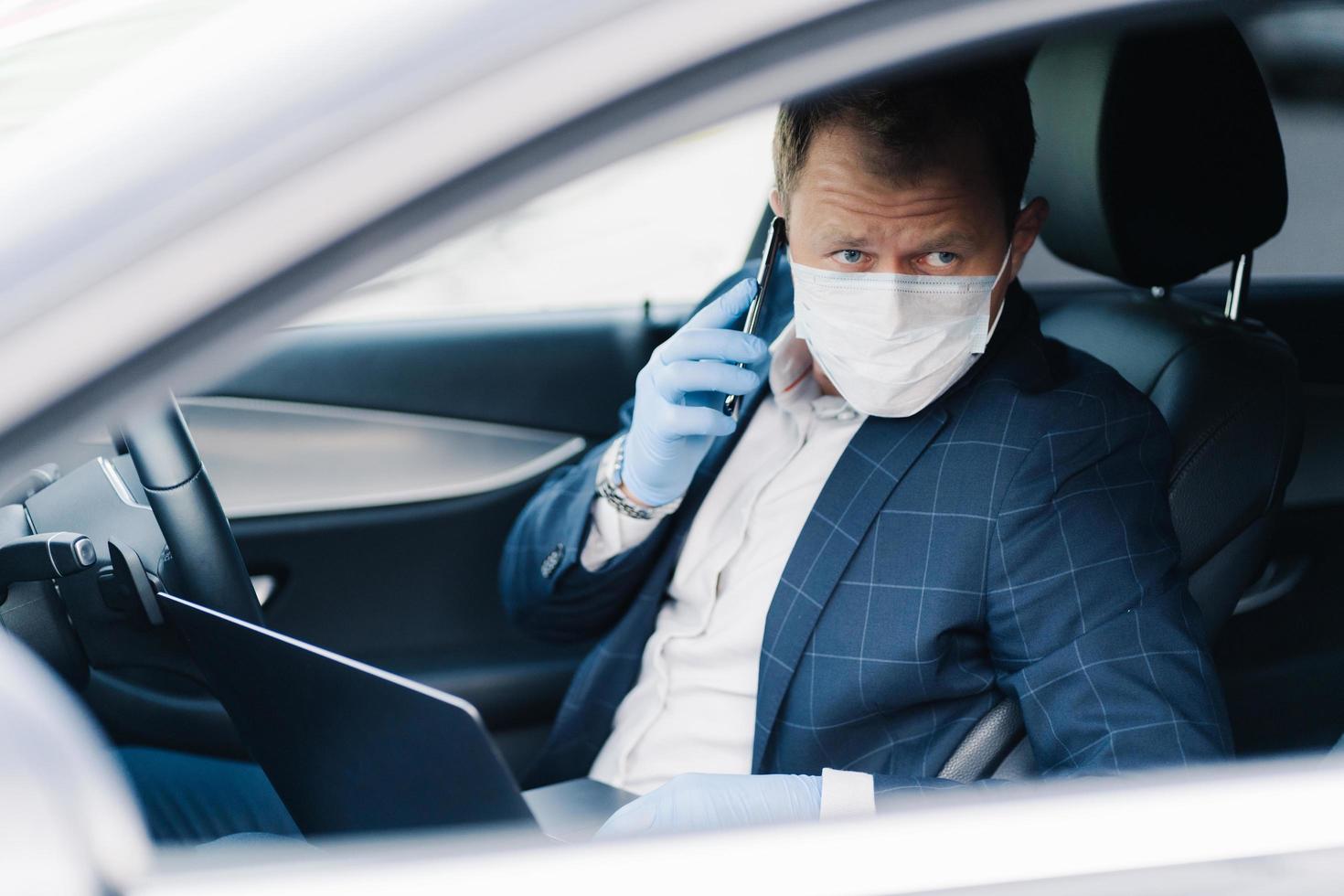 Serious prosperous entrepreneur poses in own transport, has telephone conversation, uses modern technologies to be always in touch, wears medical mask and gloves during quarantine and pandemic photo