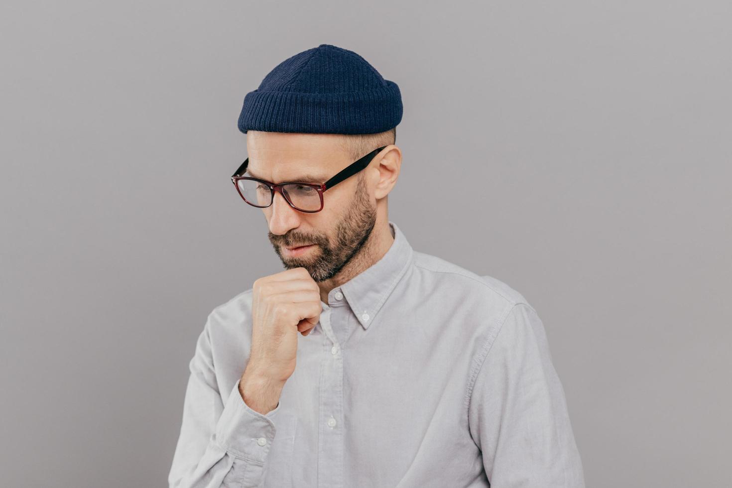 Indoor shot of concentrated man holds chin and focused down, being in deep in thoughts, thinks about innovative idea for his research, has clever expression, wears glasses and white formal shirt photo