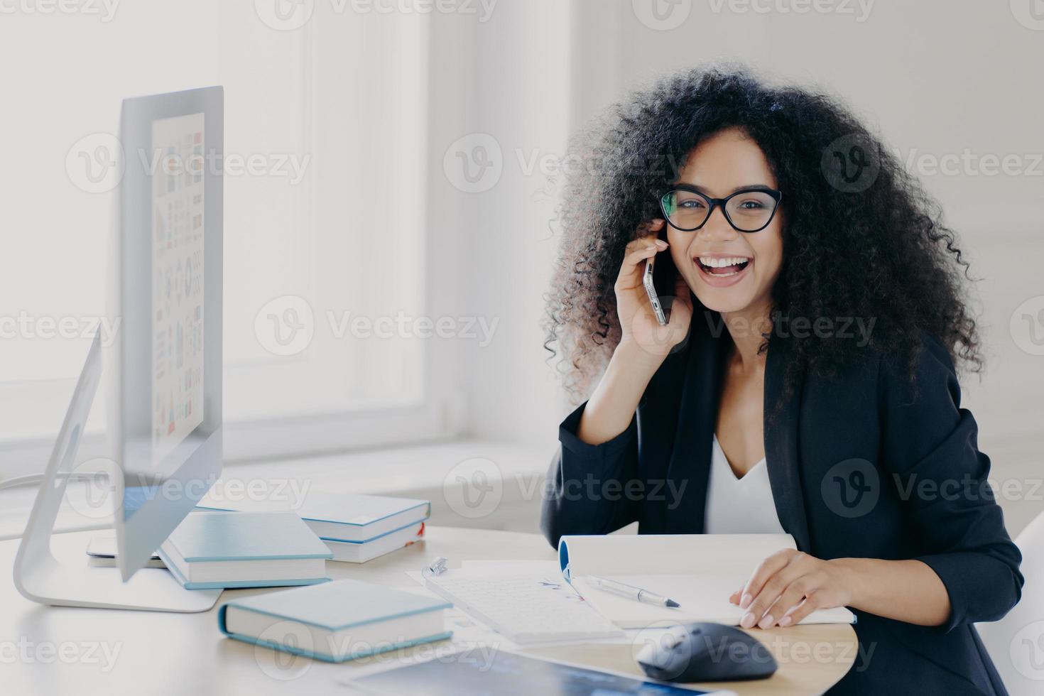 Optimistic female Afro American entrepreneur with curly hairstyle, holds mobile phone, enjoys telephone conversation with colleague, works on computer, poses at workplace with notepad and books photo