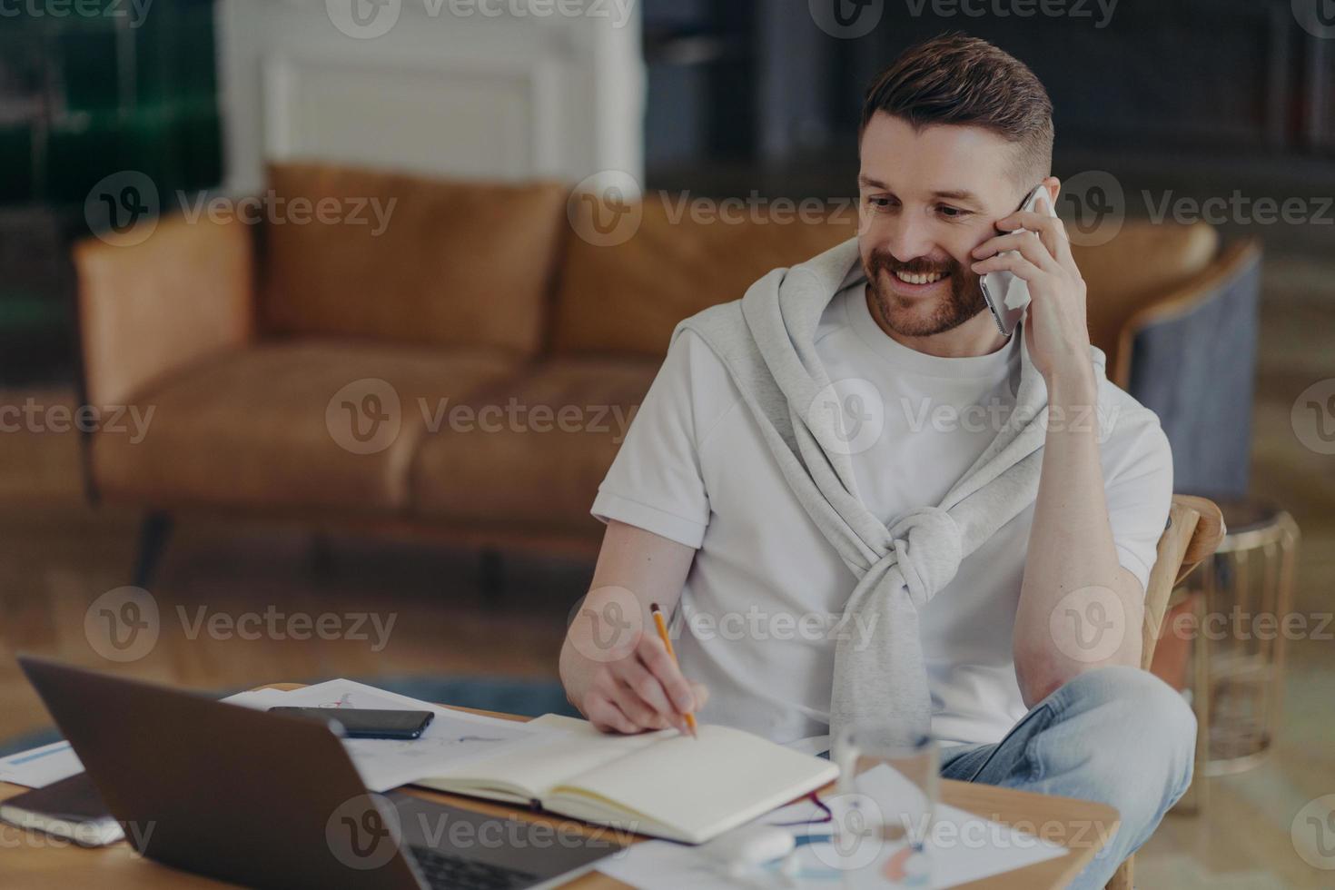 Cheerful man freelancer makes telephone call writes down information in notepad makes notes in planner looks attentively at laptop screen poses against home interior at desktop. Technology user photo