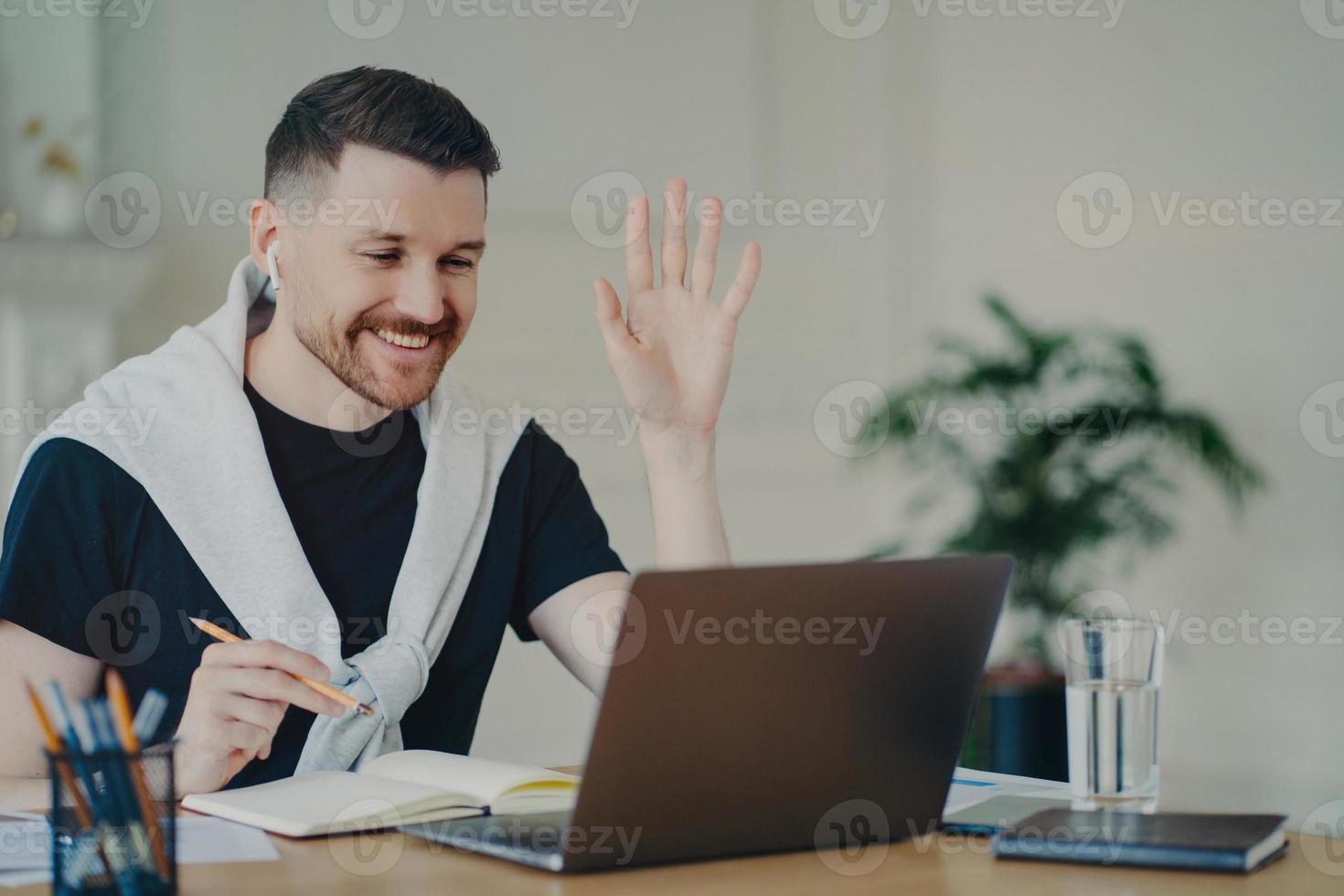 Cheerful bearded man has video conference online waves palm in laptop computer uses wireless earphones poses at coworking space dressed casually writes down information in diary smiles gladfully photo