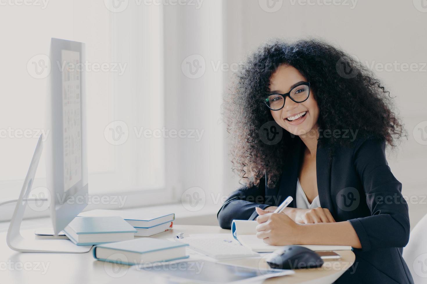 Cheerful female student writes down necessary information in notebook, makes notes, has glad expression, wears transparent glasses and black formal wear, sits at desktop with books and computer photo