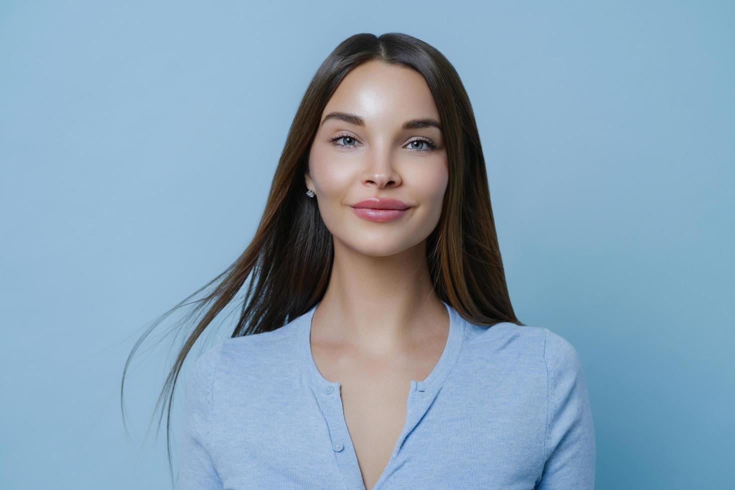 Indoor portrait of pretty woman feels happy and carefree during day off, has long straight hair, wears blue jumper, enjoys positive conversation, stands indoor. Women, lifestyle and well being concept photo